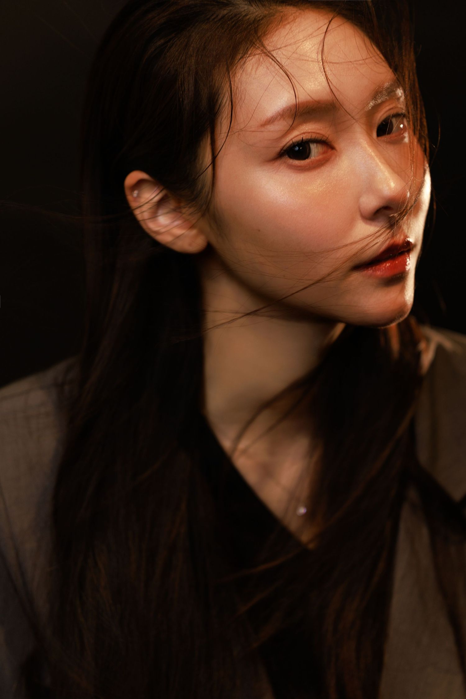 Young Asian woman with long dark hair in dramatic lighting, wearing a gray jacket, gazing thoughtfully to the side.