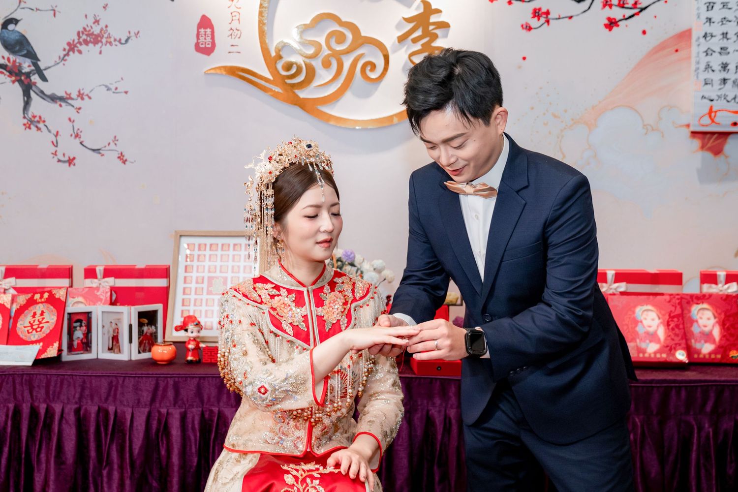 Bride in red and gold traditional Chinese wedding dress with ornate headpiece exchanges rings with groom in navy suit during tea ceremony.