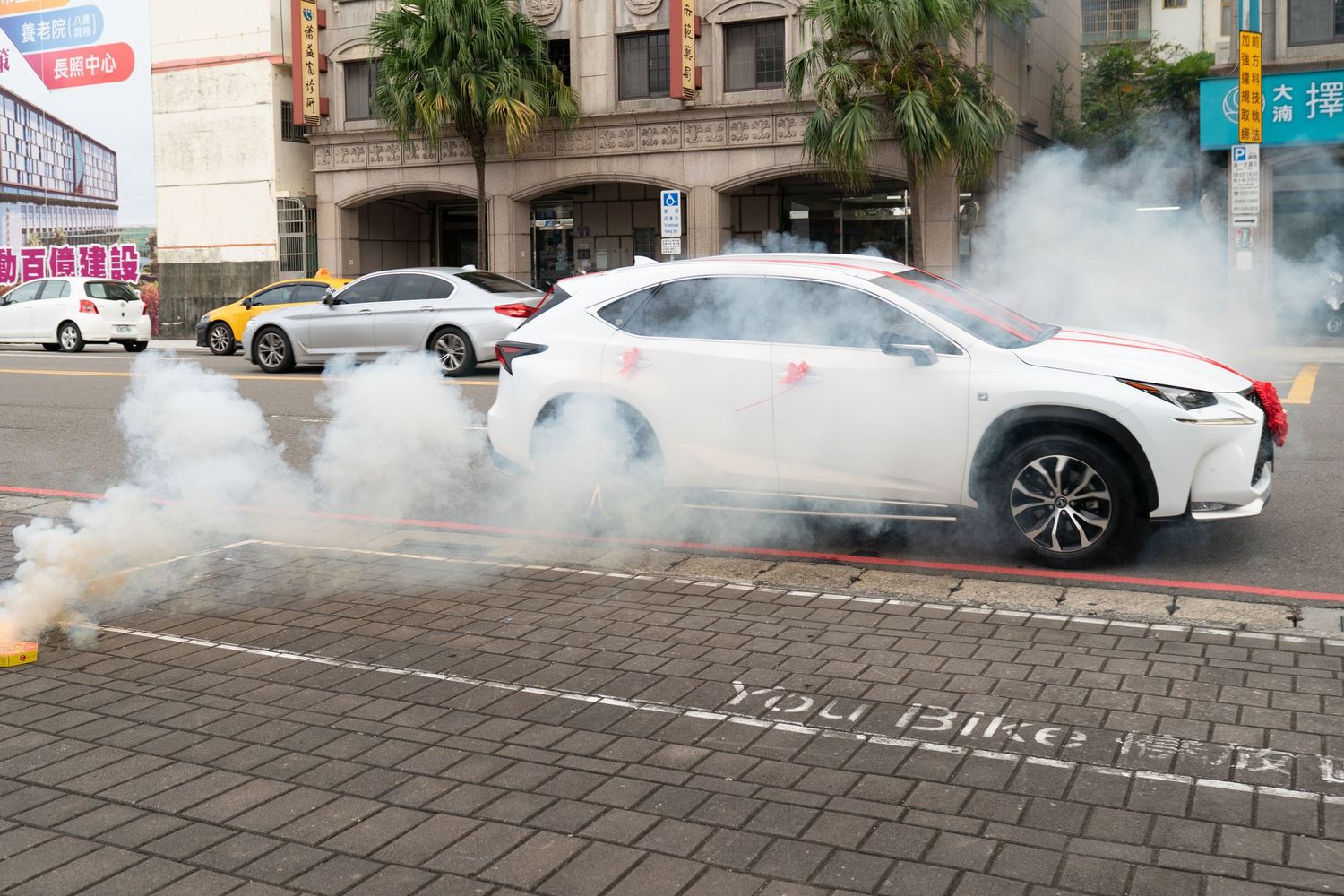 White Lexus SUV performing a burnout on a city street, creating large smoke clouds near a bike lane marking.