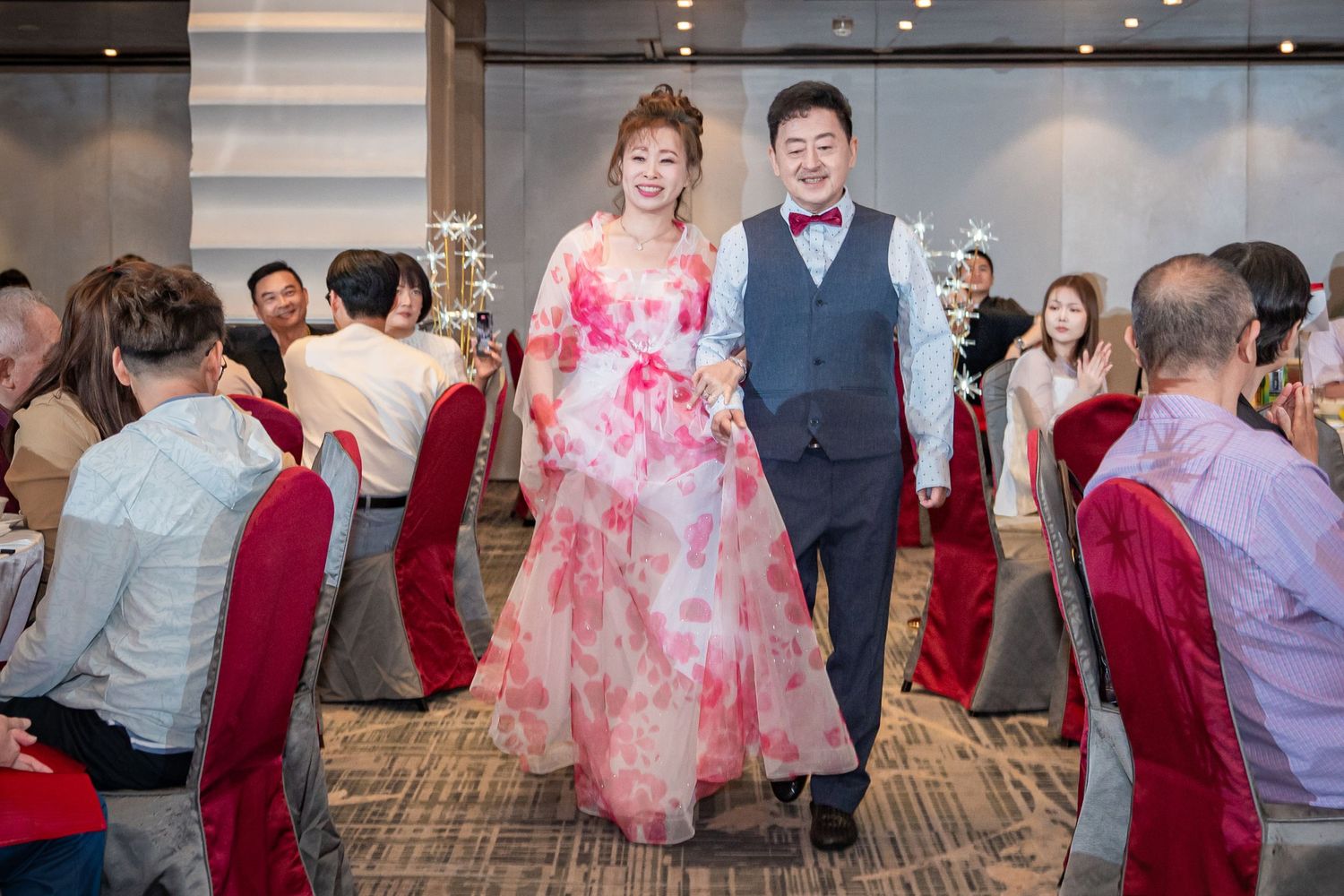 Couple walks arm-in-arm through a wedding banquet hall, bride in floral pink gown, groom in gray suit with bow tie.