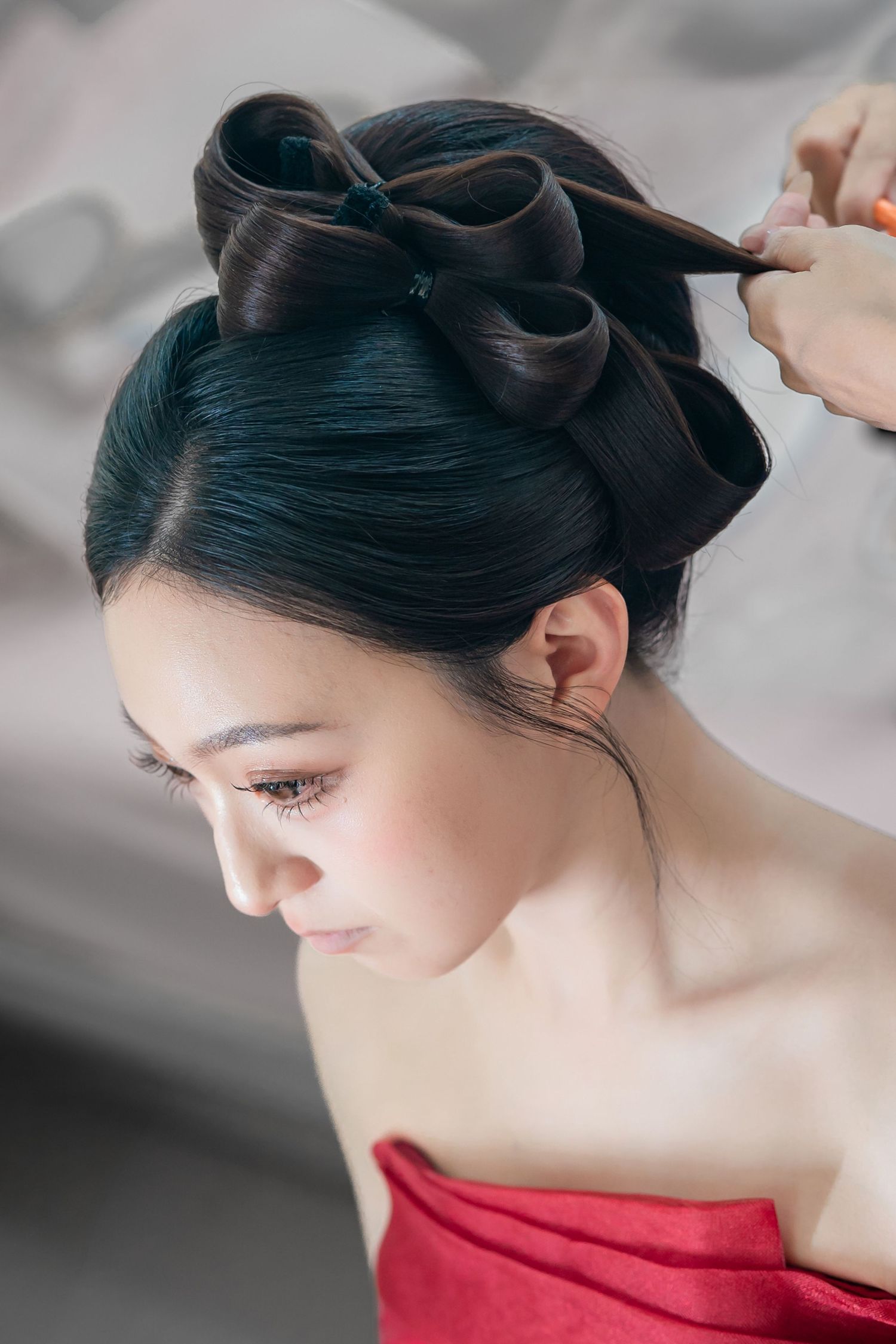 A hairstylist creates an elegant bow-shaped updo on a young Asian woman wearing a red dress.