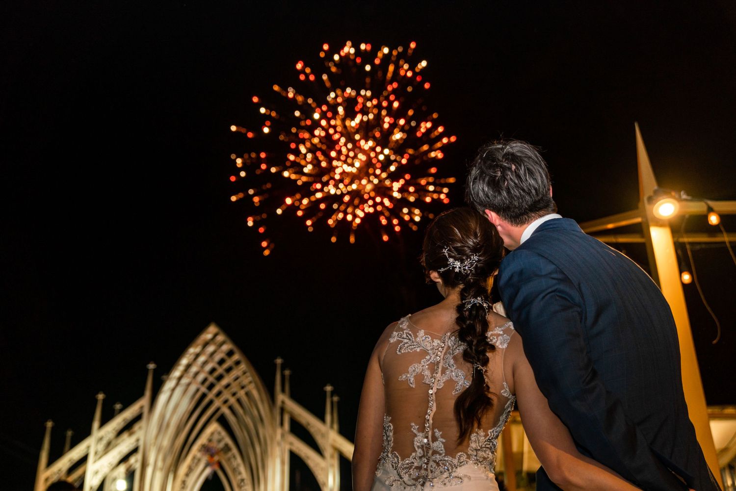 A couple watches fireworks burst in the night sky above an illuminated gothic-style building during a celebration.