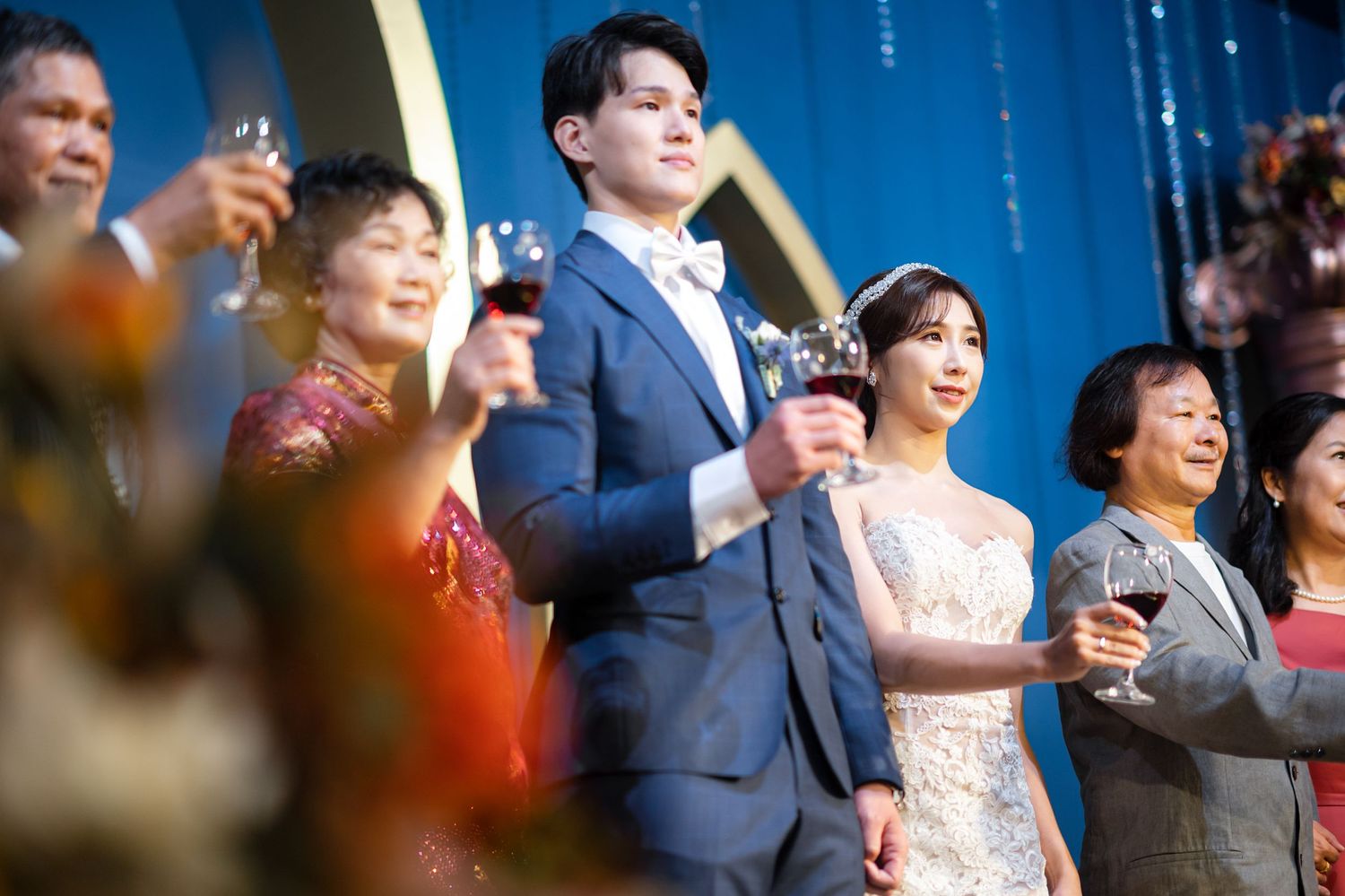 Wedding couple and guests raise glasses for a celebratory toast against a blue curtain backdrop.