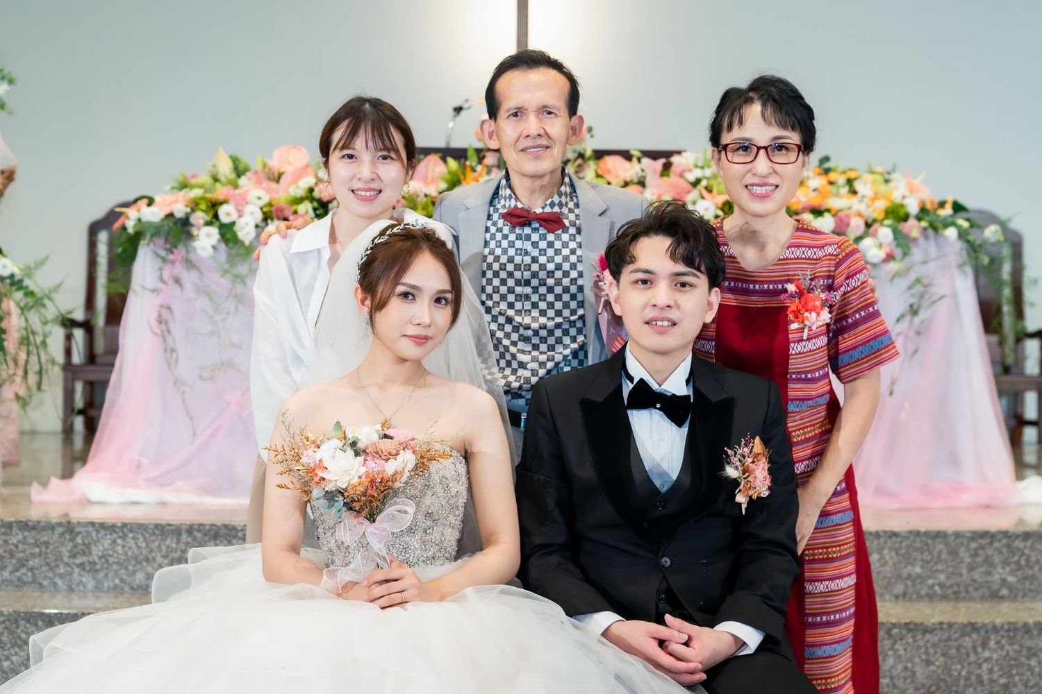 Wedding portrait with guests in traditional and modern attire against floral backdrop.