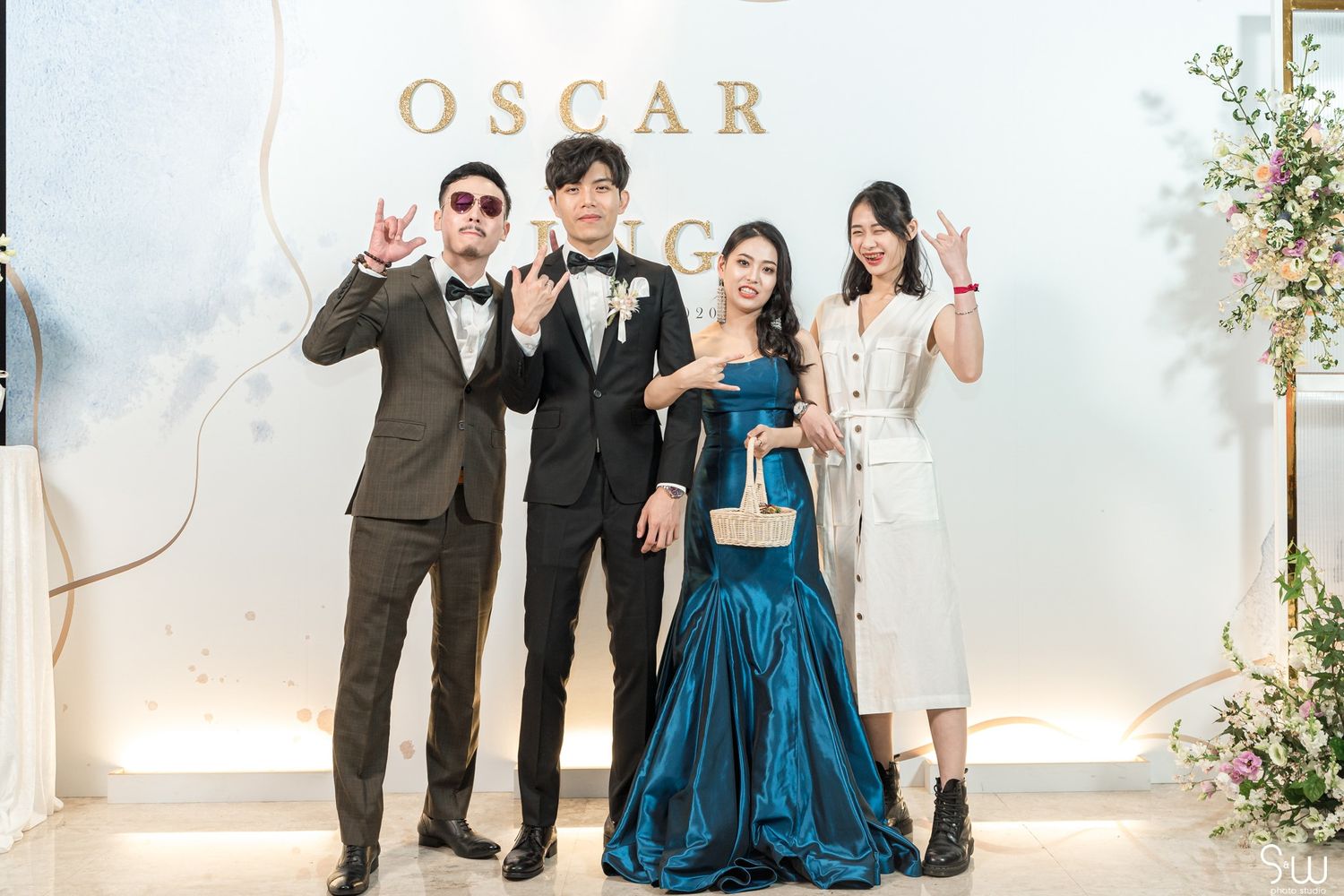 Wedding party poses in formal attire against a white floral backdrop with 'OSCAR' signage.