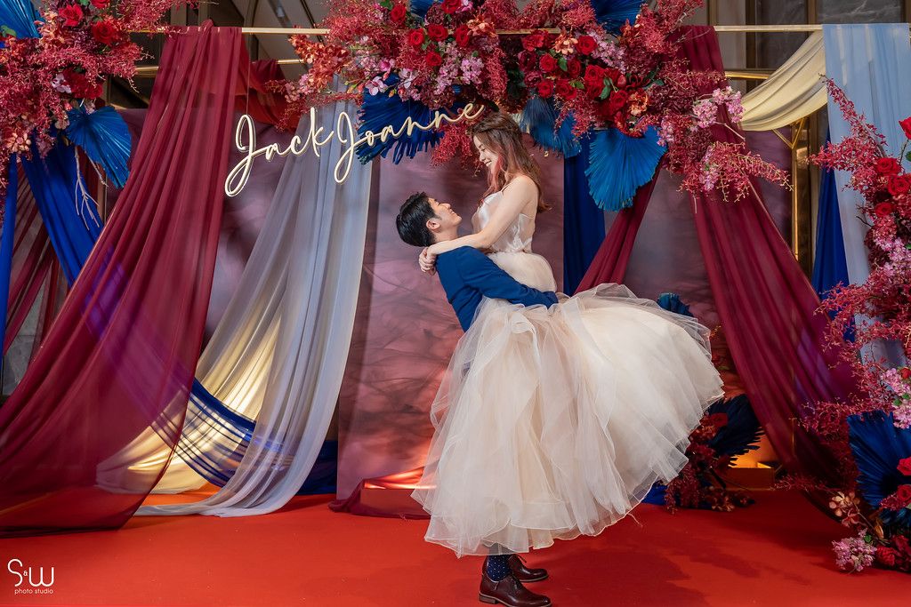 Romantic dip pose during wedding celebration against red and blue floral backdrop.