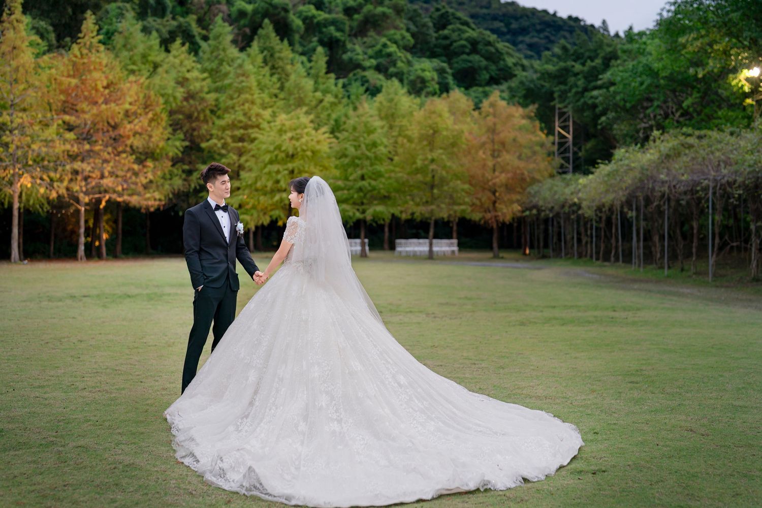 A couple in wedding attire poses together in a lush outdoor garden setting with trees and greenery in the background.