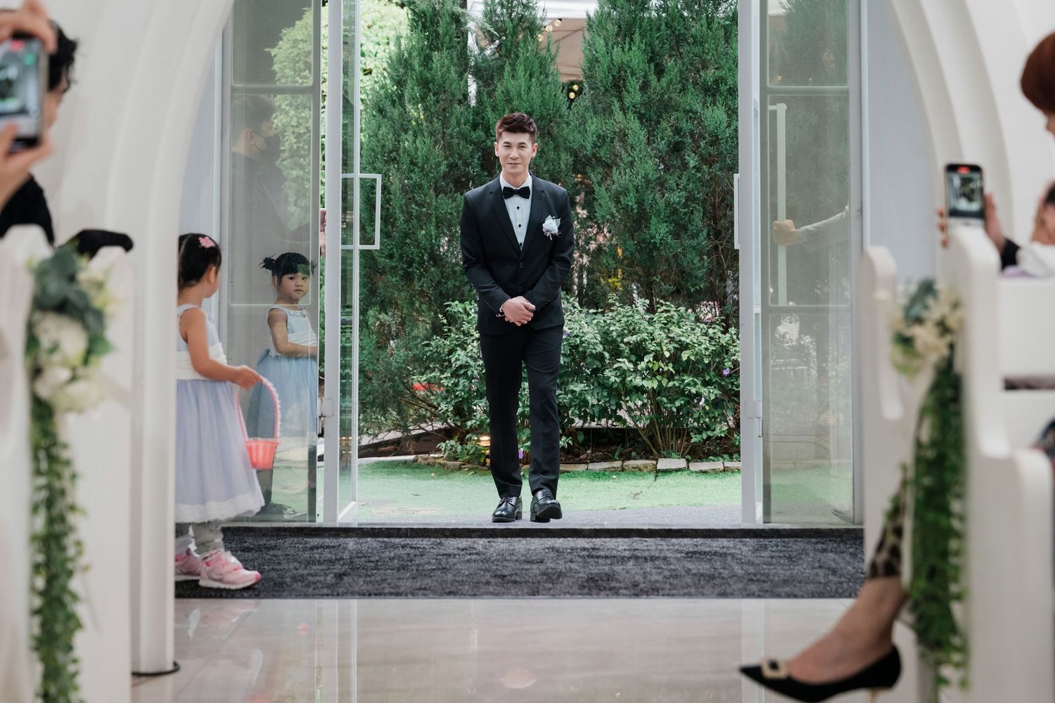 A wedding party poses in an elegant glass-enclosed walkway surrounded by lush greenery and modern architecture.