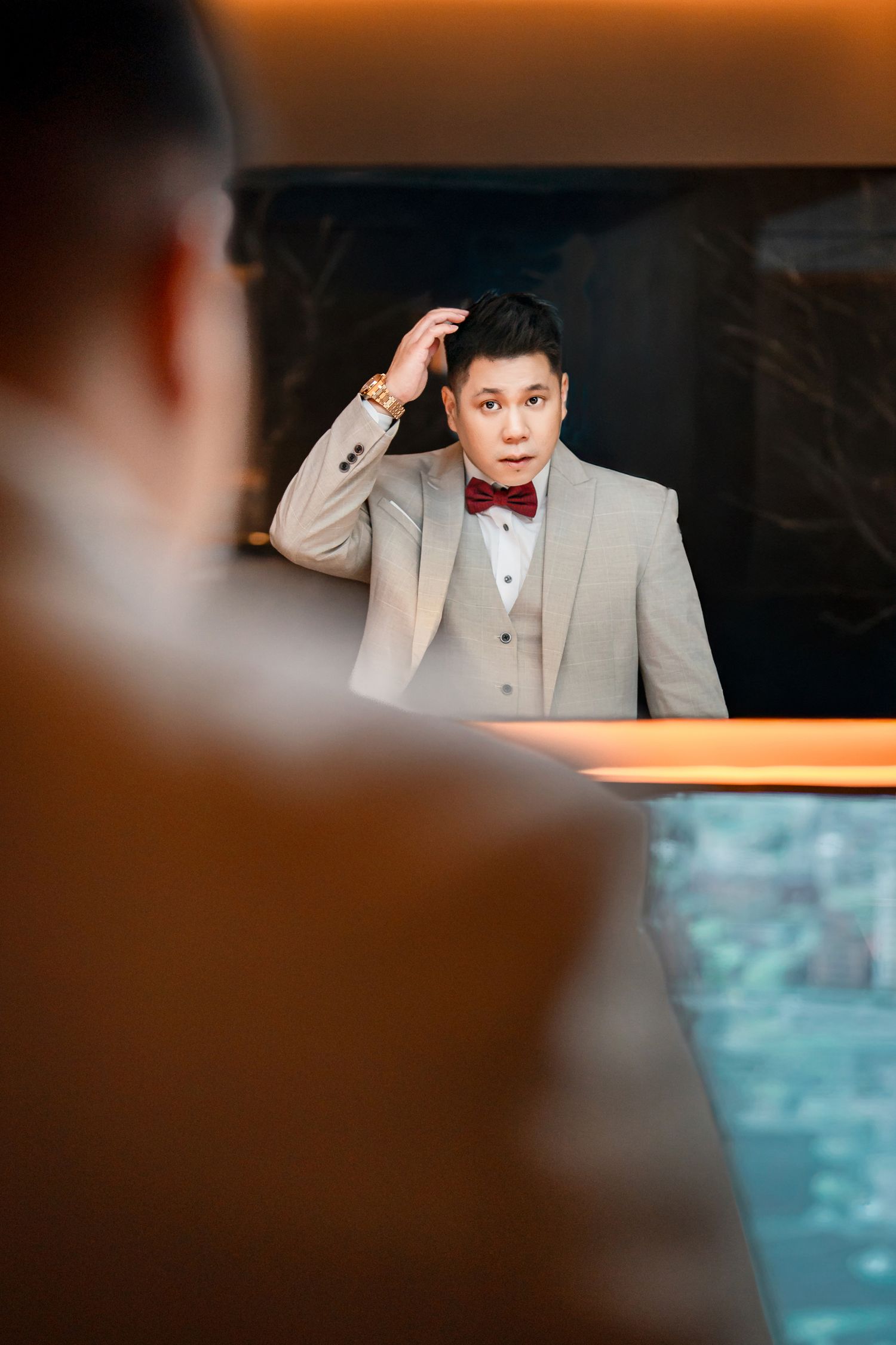 A person in a light gray suit and red bow tie adjusts their collar while posing near a reflective surface.