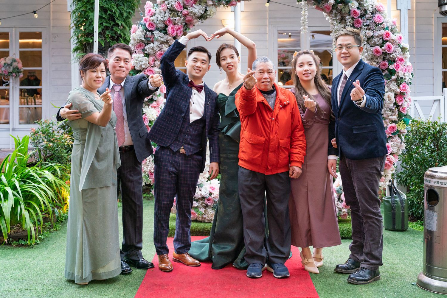 A group stands together on a red carpet in front of a floral arch at an outdoor event venue.