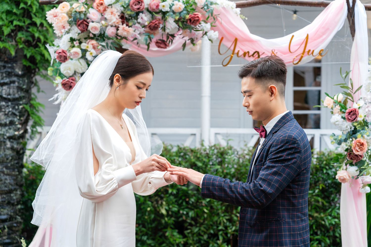 Two people exchange rings during a romantic outdoor wedding ceremony under a floral arch with pink roses.