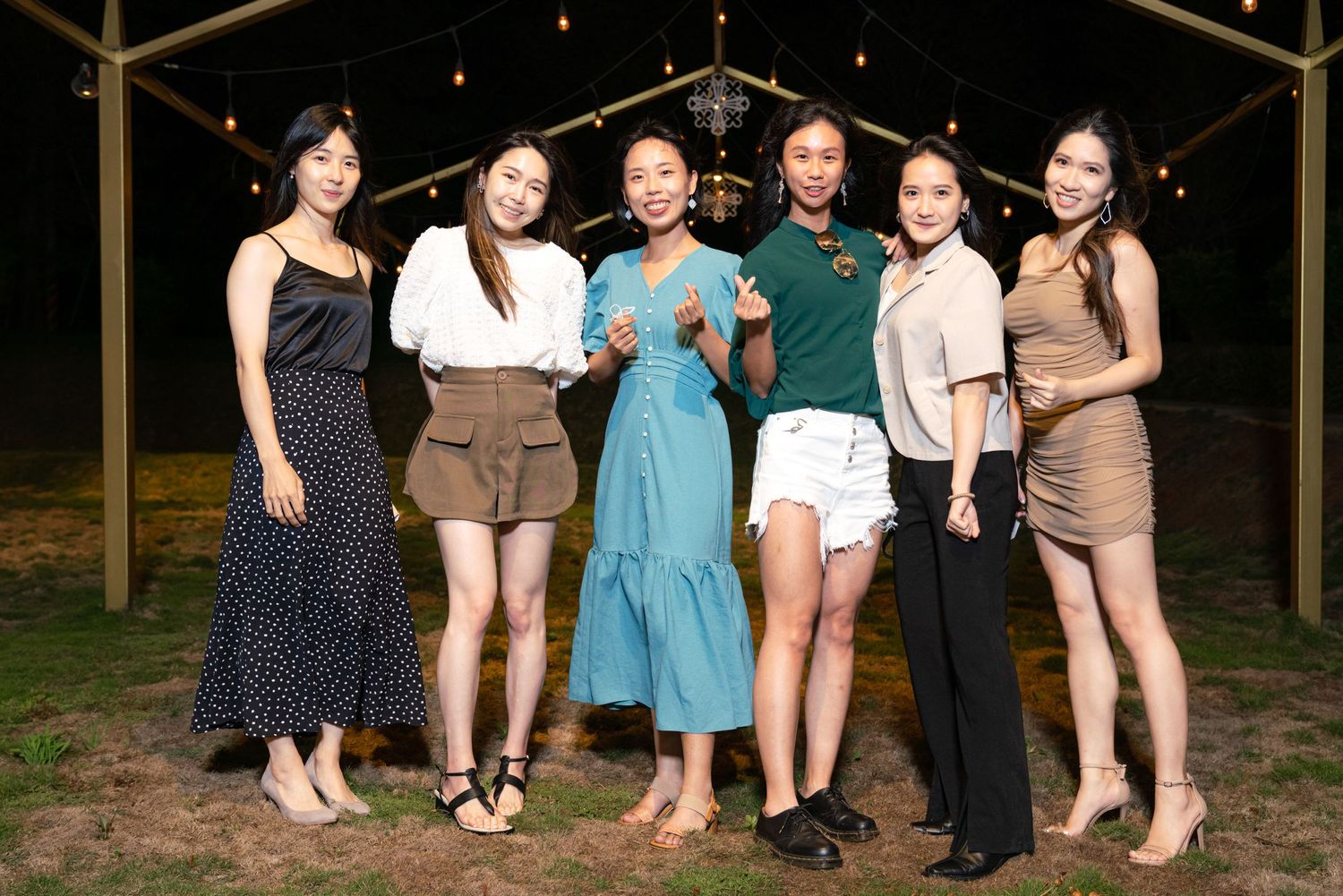 A group of six friends pose together outdoors at night under string lights wearing casual summer attire.