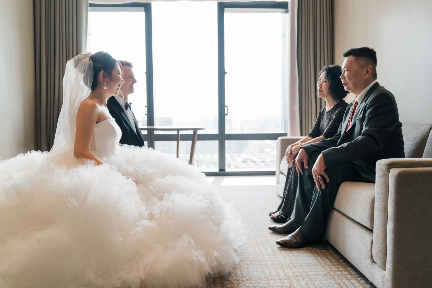 A bride in a white wedding gown sits on a couch next to a large window in an elegant room.