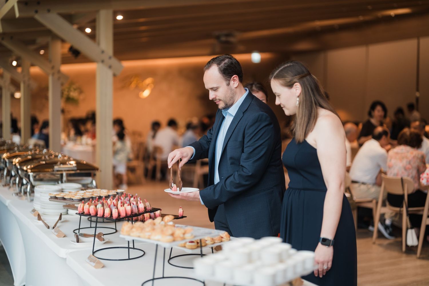 Wedding reception guests browse an elegant dessert display table featuring pink macarons and treats at an indoor venue.