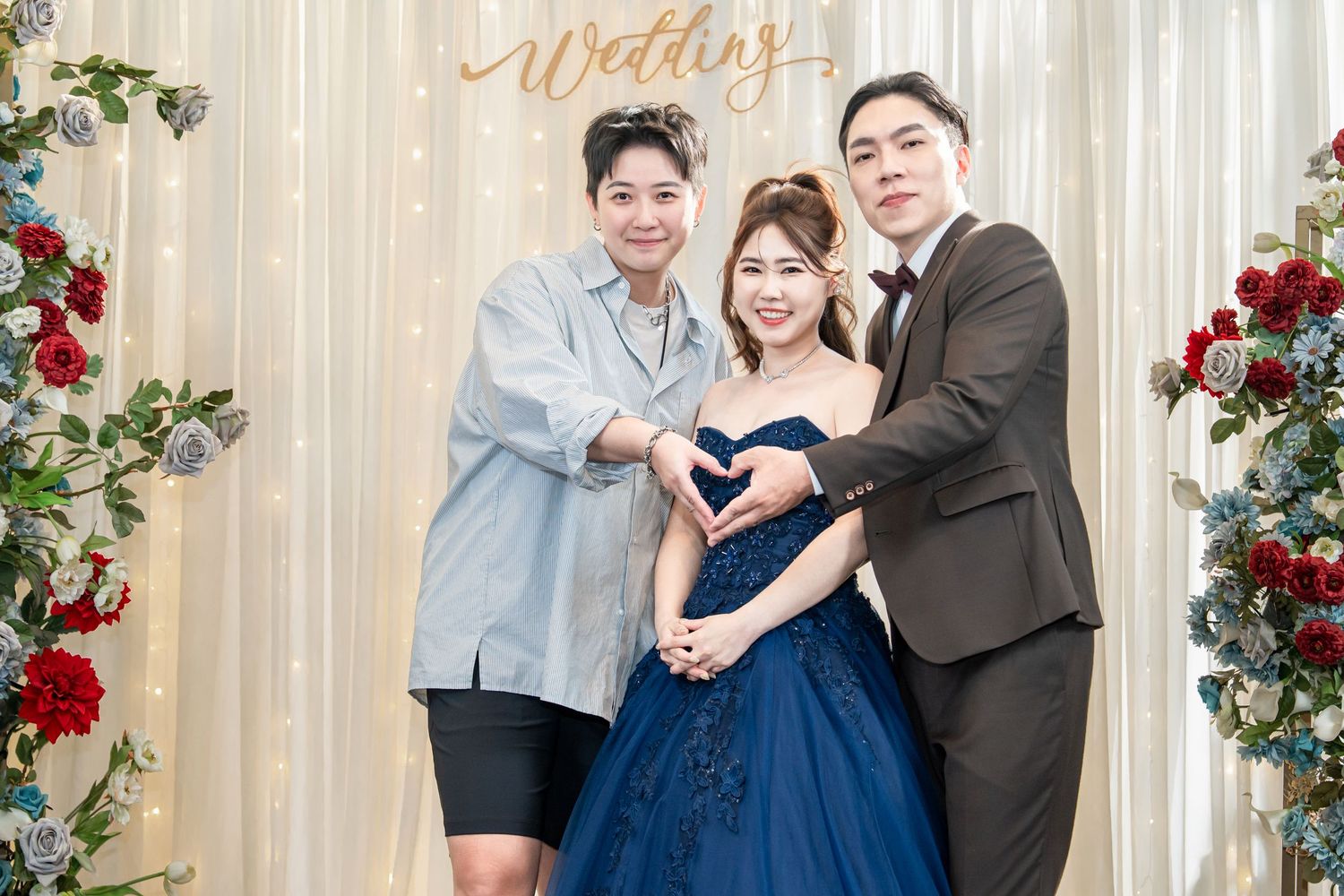 Three people make heart gestures while posing at an elegant wedding backdrop with floral decor.