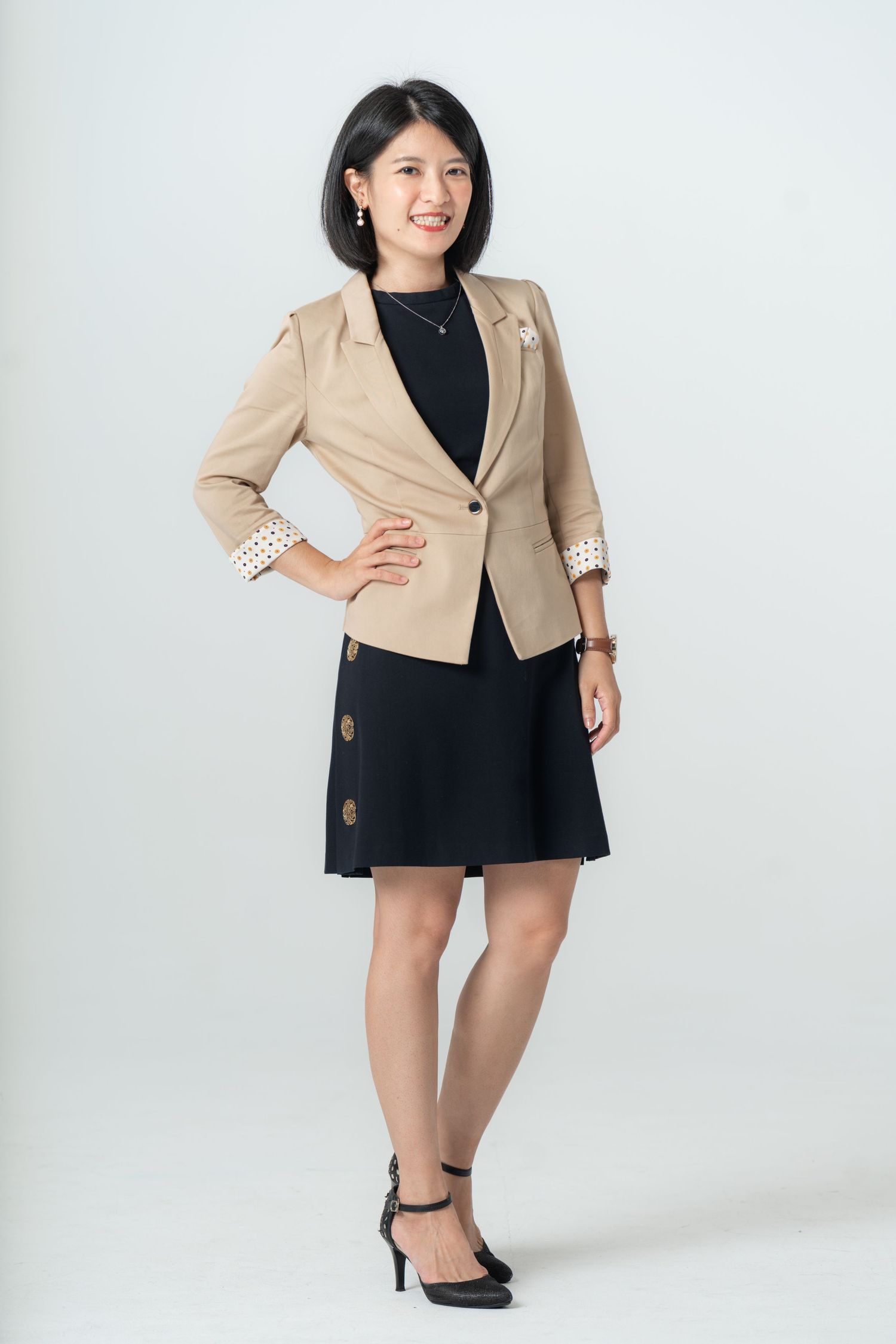 Professional model wearing a beige blazer with lace trim and black dress poses confidently in a studio setting.