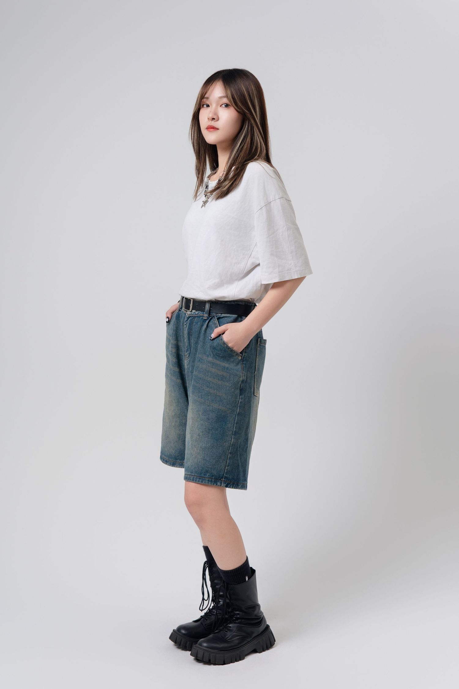 Fashion model wearing white t-shirt, denim shorts and chunky black boots in studio setting against grey backdrop.