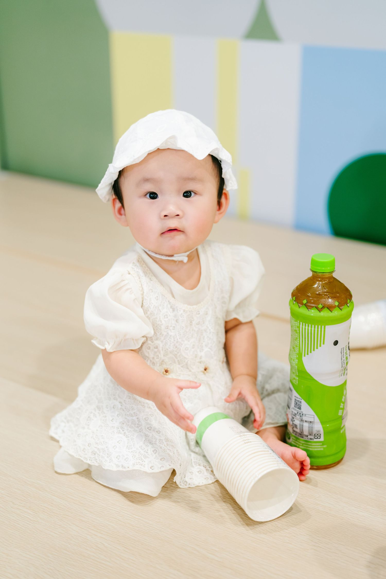 A baby in a white dress sits on a hardwood floor with bottles of green drinks beside them.