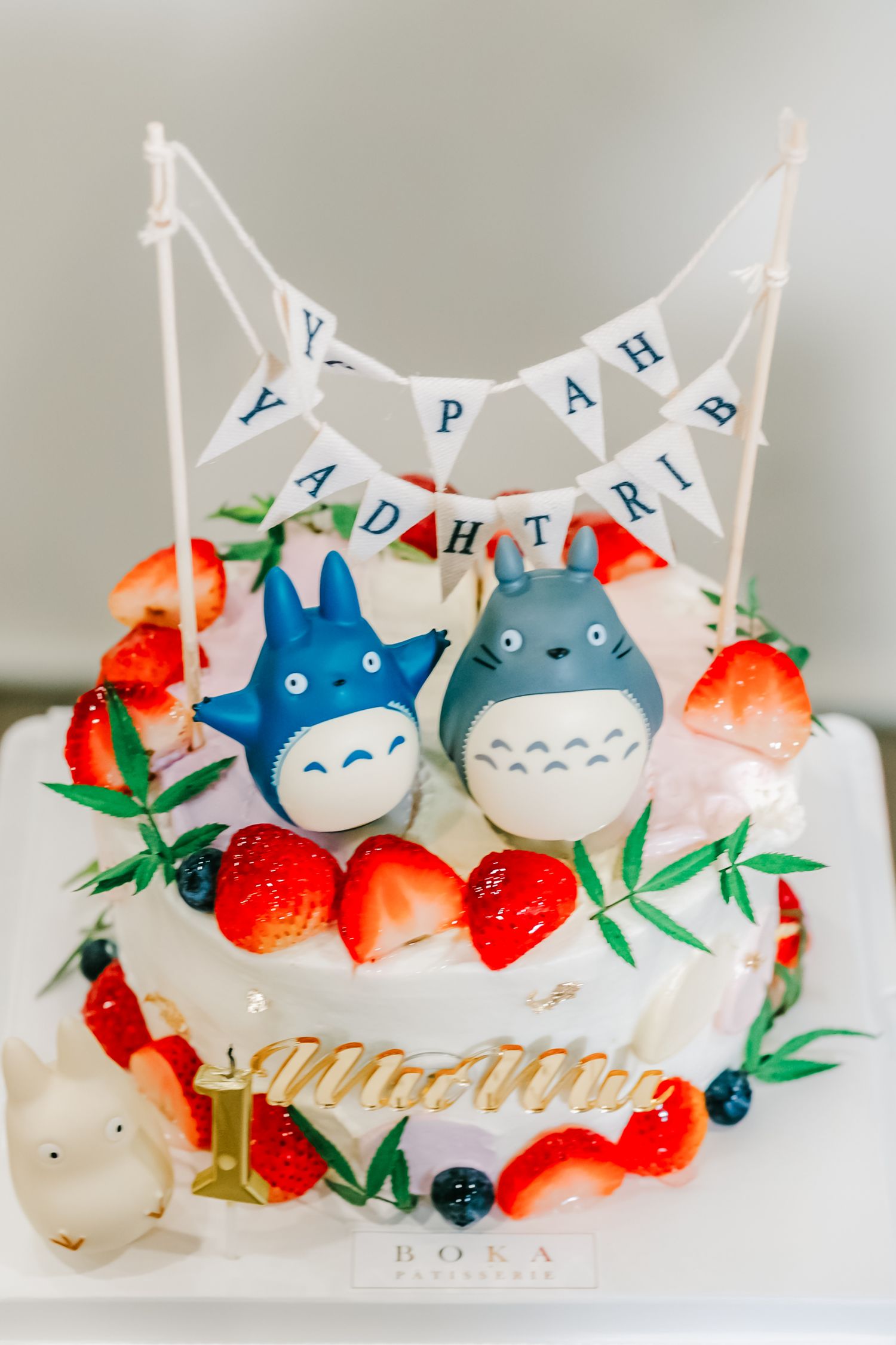 White birthday cake decorated with Totoro characters, fresh strawberries, blueberries and a 'Happy Birthday' banner on top.