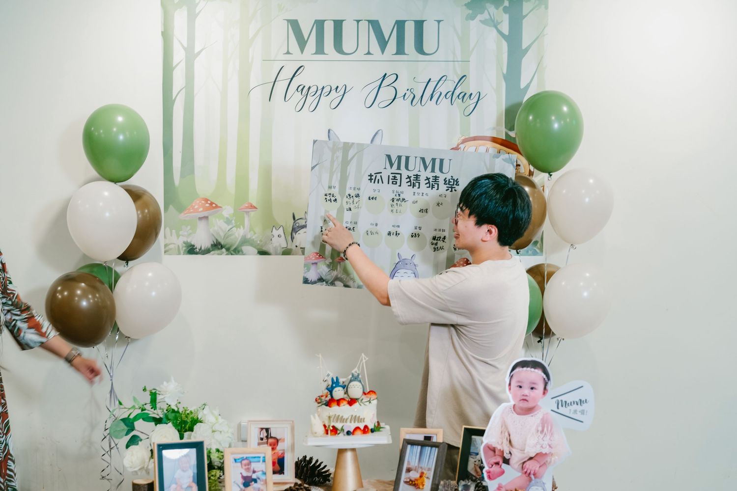 A birthday party setup with green and white balloons, forest-themed backdrop, and someone decorating by the wall.