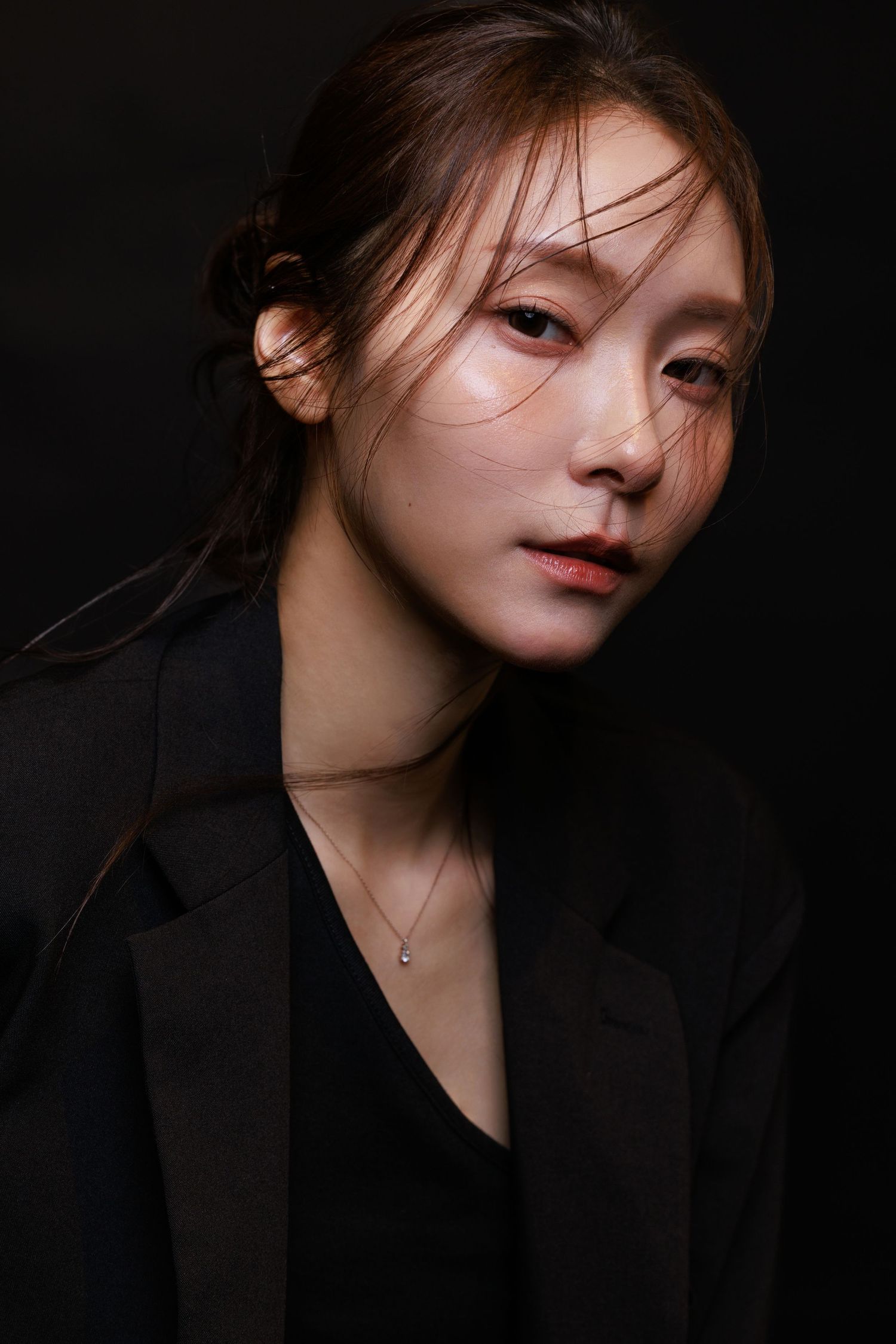 Young Asian woman in black blazer with delicate necklace poses against dark background with dramatic lighting.