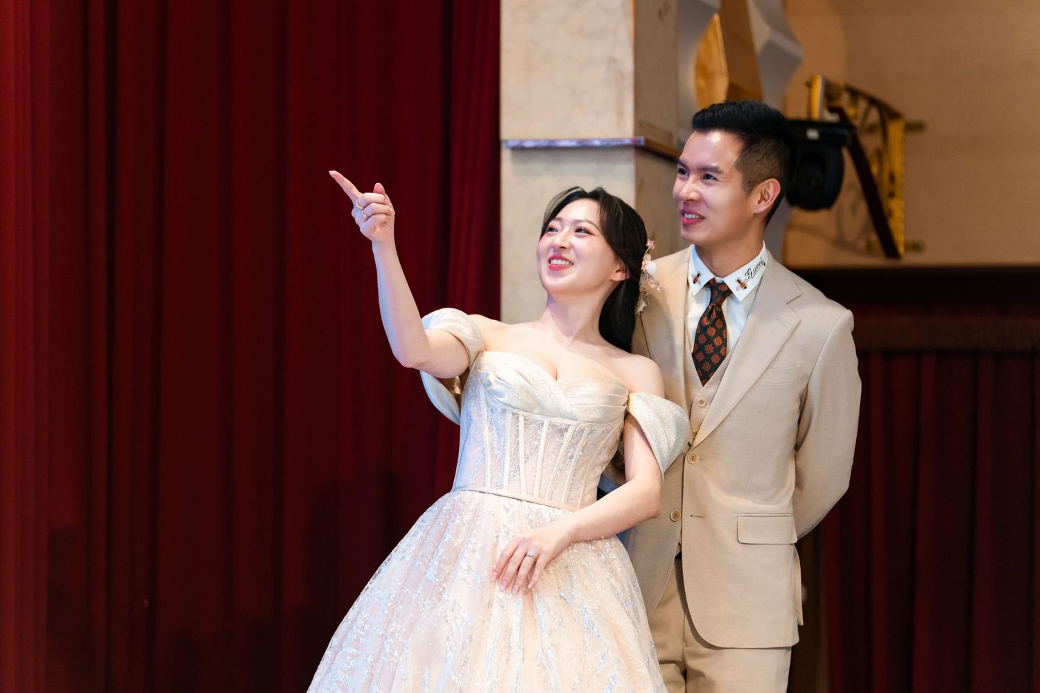 Elegant couple in formal attire, woman in white ball gown pointing, man in beige suit, on stage.