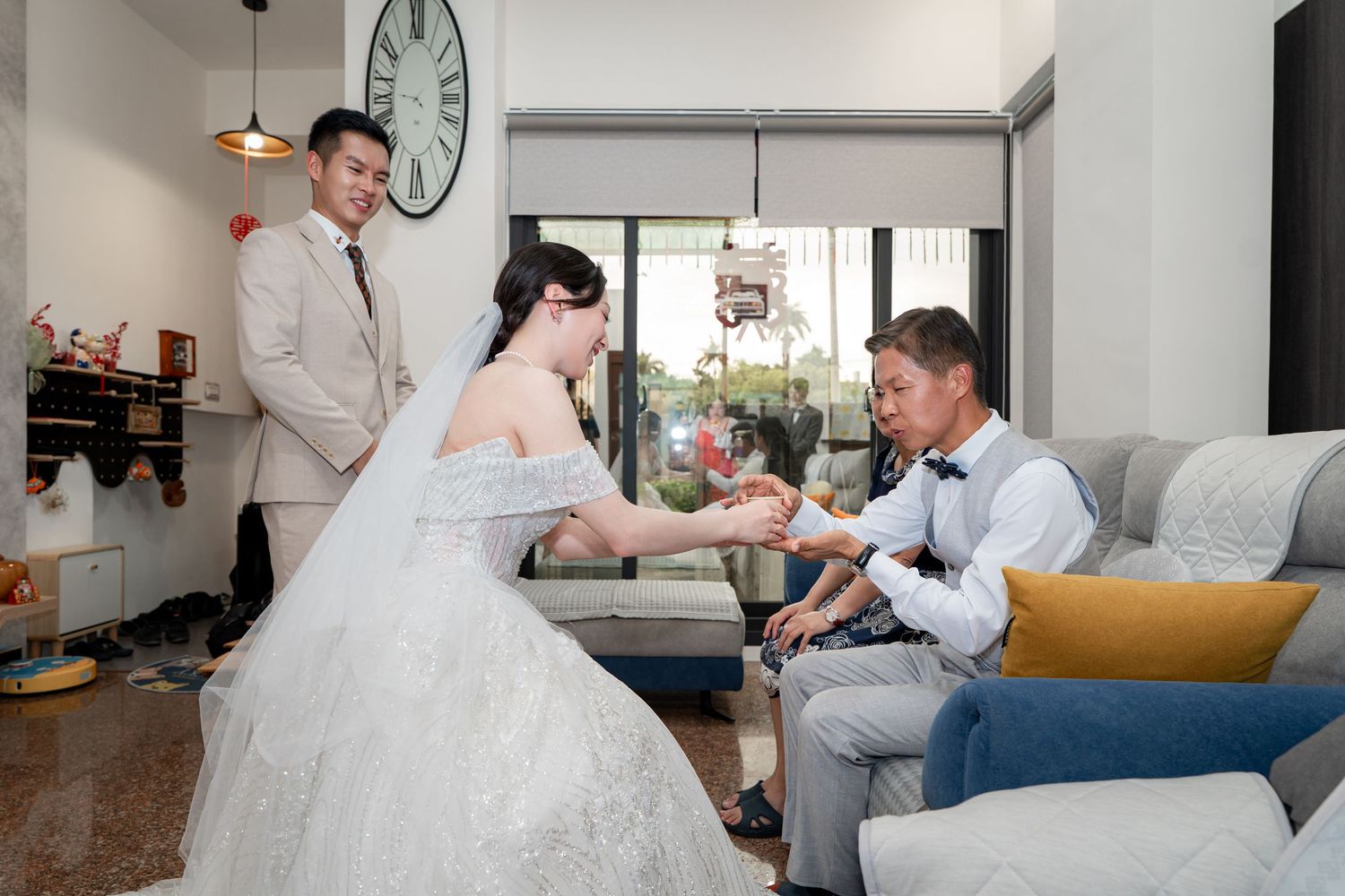 Bride in white lace gown exchanges rings with groom seated on sofa as groomsman watches in modern living room.