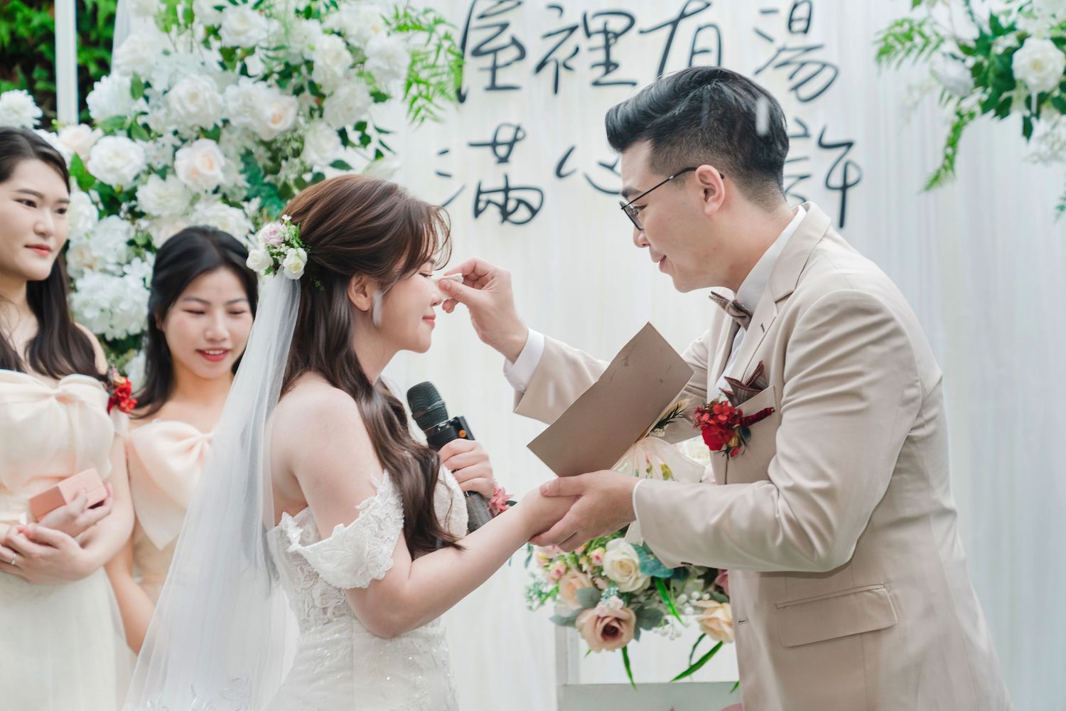 A joyful couple celebrates their wedding ceremony under white floral decorations and calligraphy wall art.