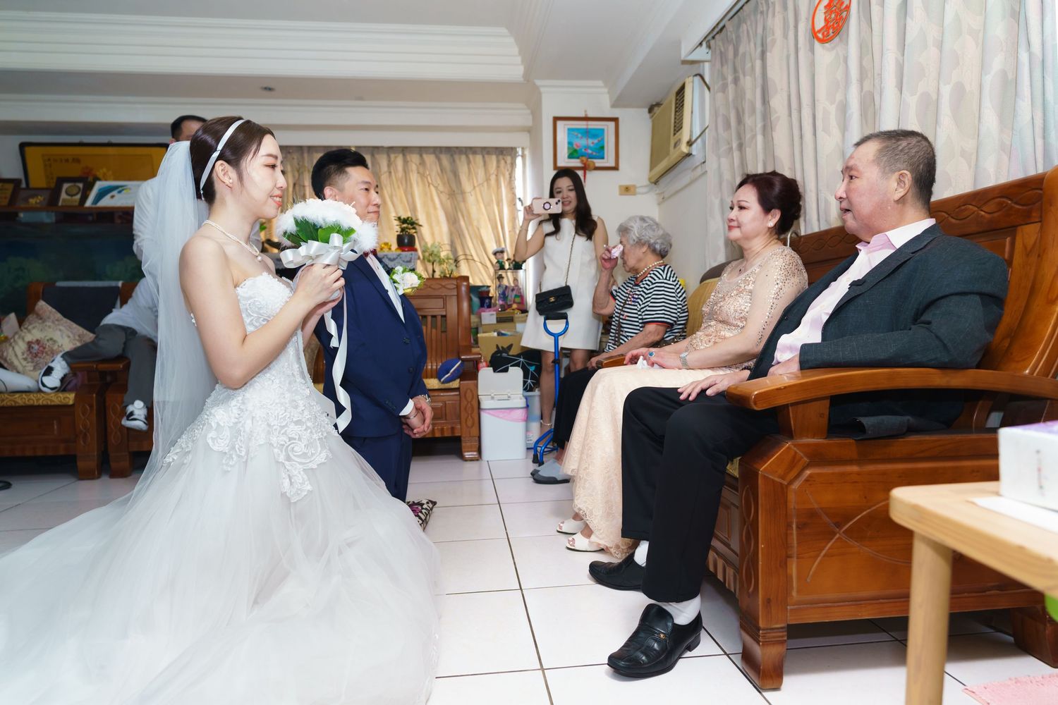 People gather in a living room with wooden furniture as someone in a white wedding dress holds a bouquet.