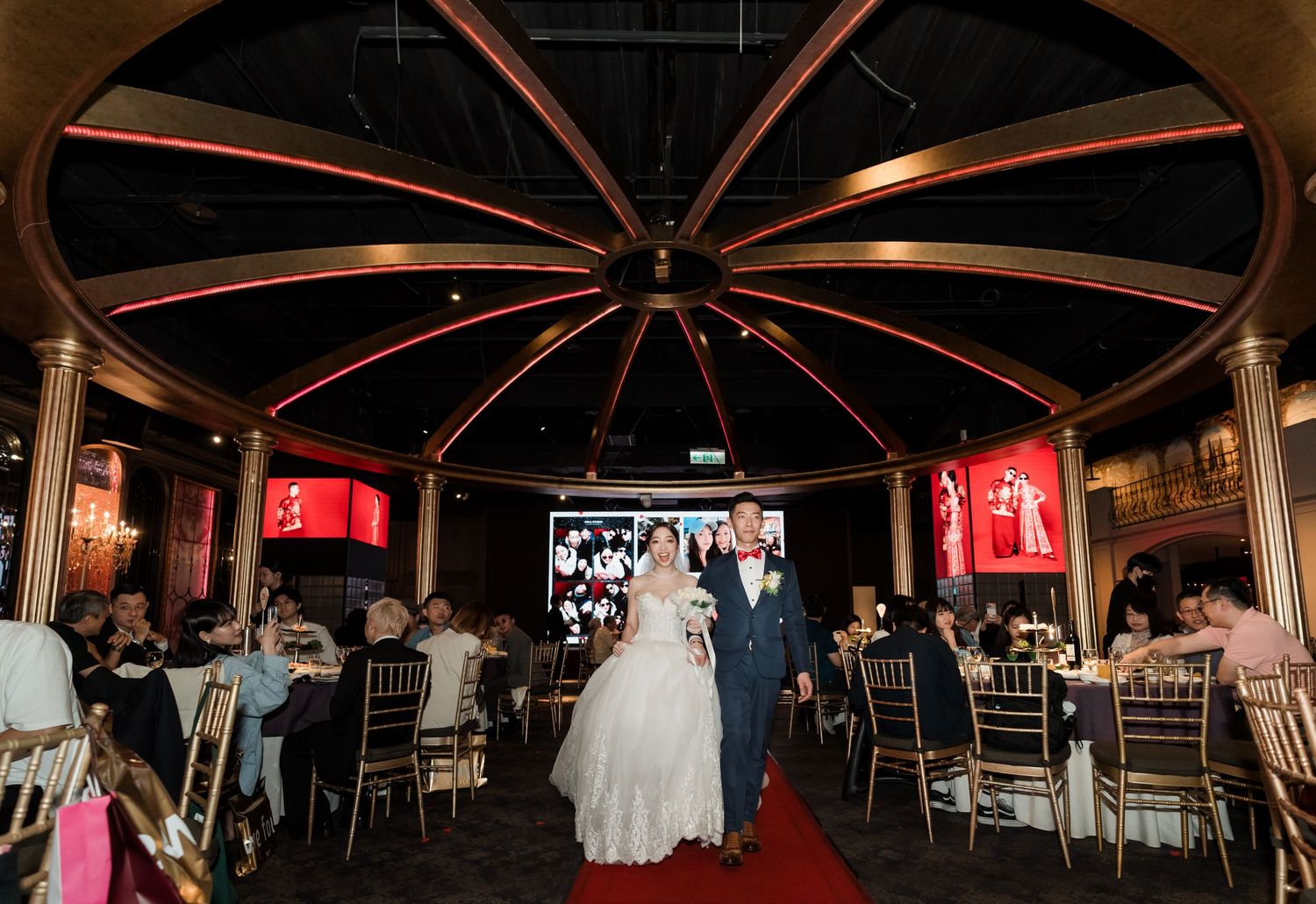 Wedding ceremony in a circular venue with red carpet aisle and illuminated ceiling beams creating a dramatic atmosphere.