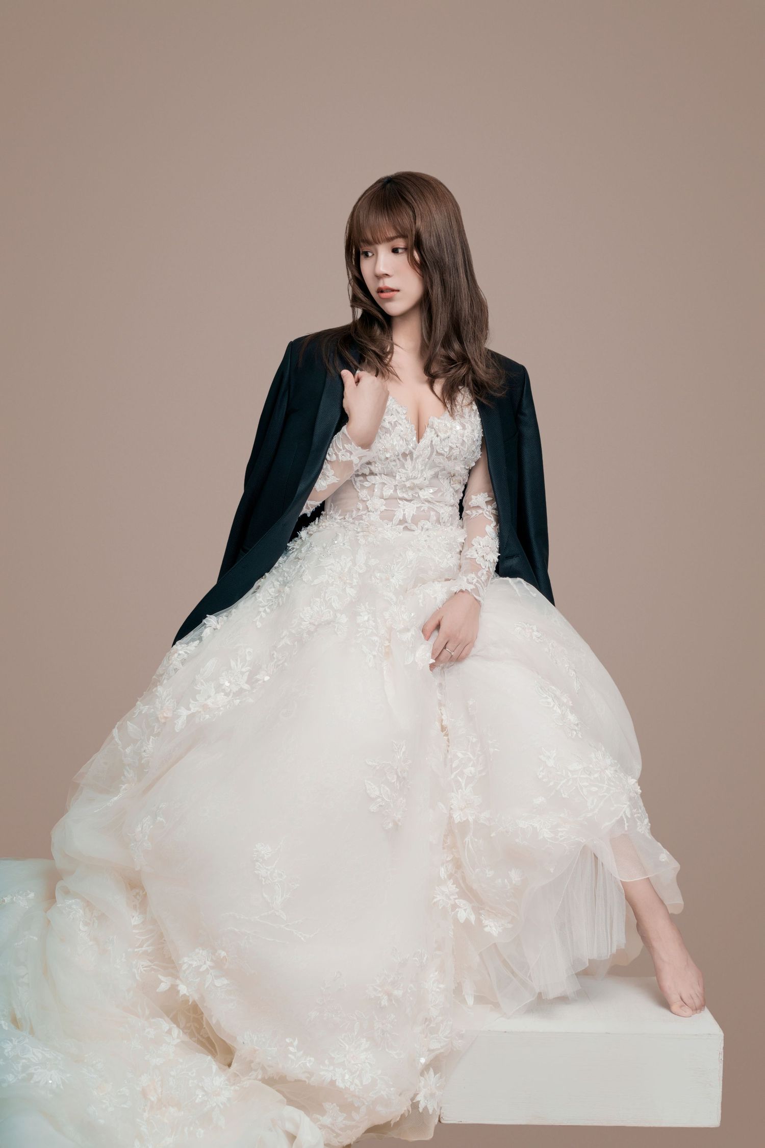 A model poses elegantly in a white lace wedding gown and dark cardigan while sitting barefoot on a white platform.