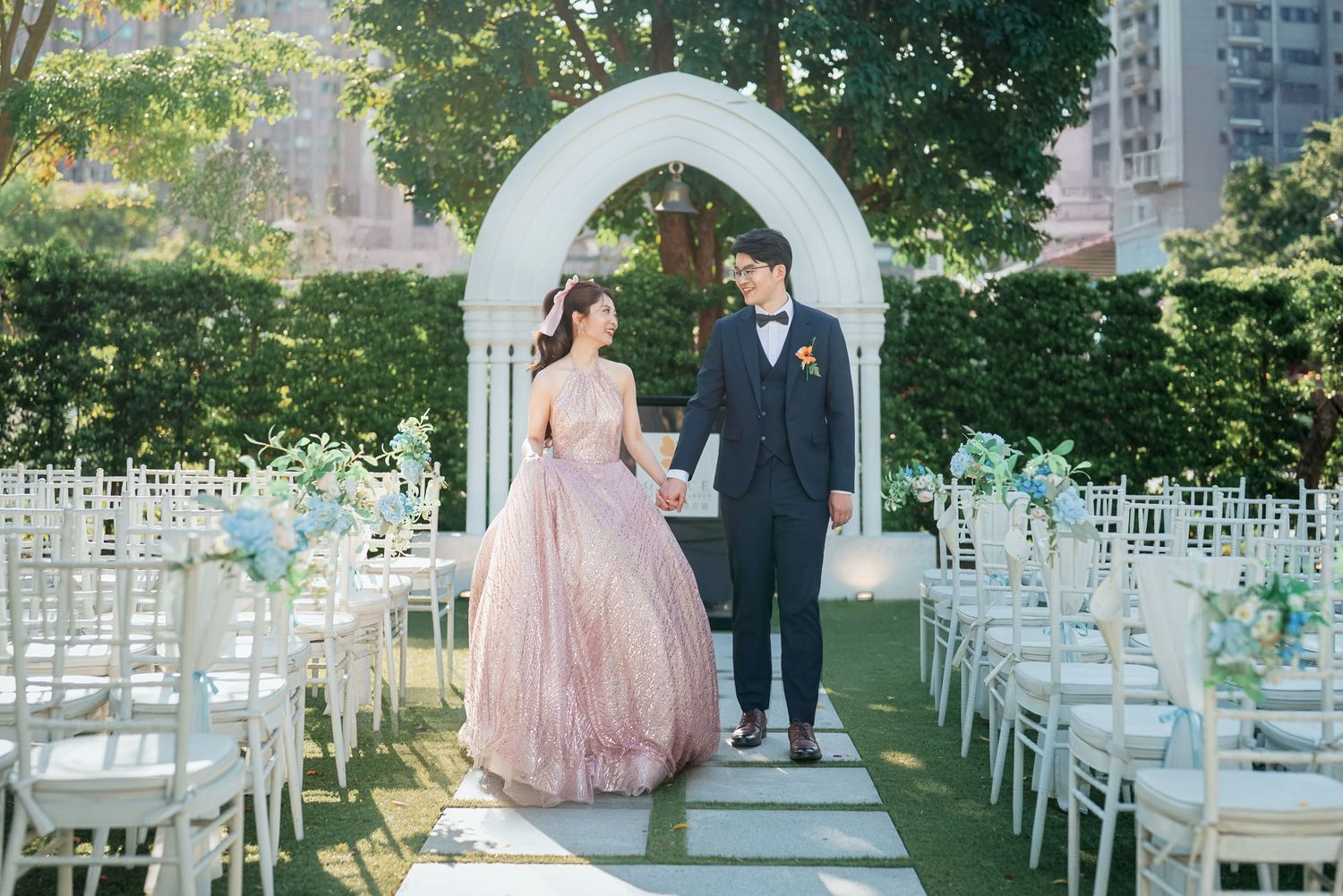 A couple stands under a white wedding arch in an outdoor garden ceremony setup with white chairs and floral decorations.