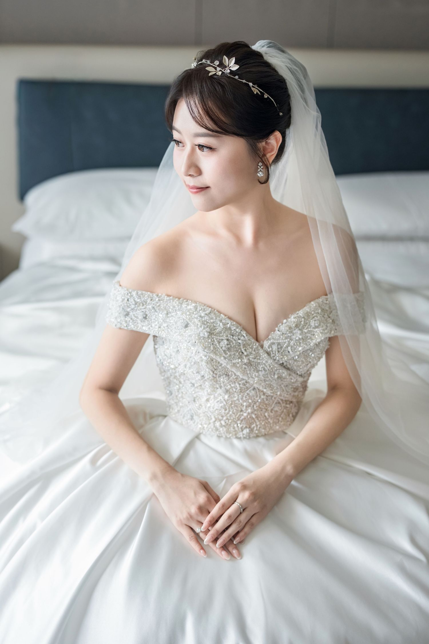 A bride in an elegant off-shoulder beaded wedding gown with tulle veil poses gracefully on white bedding.