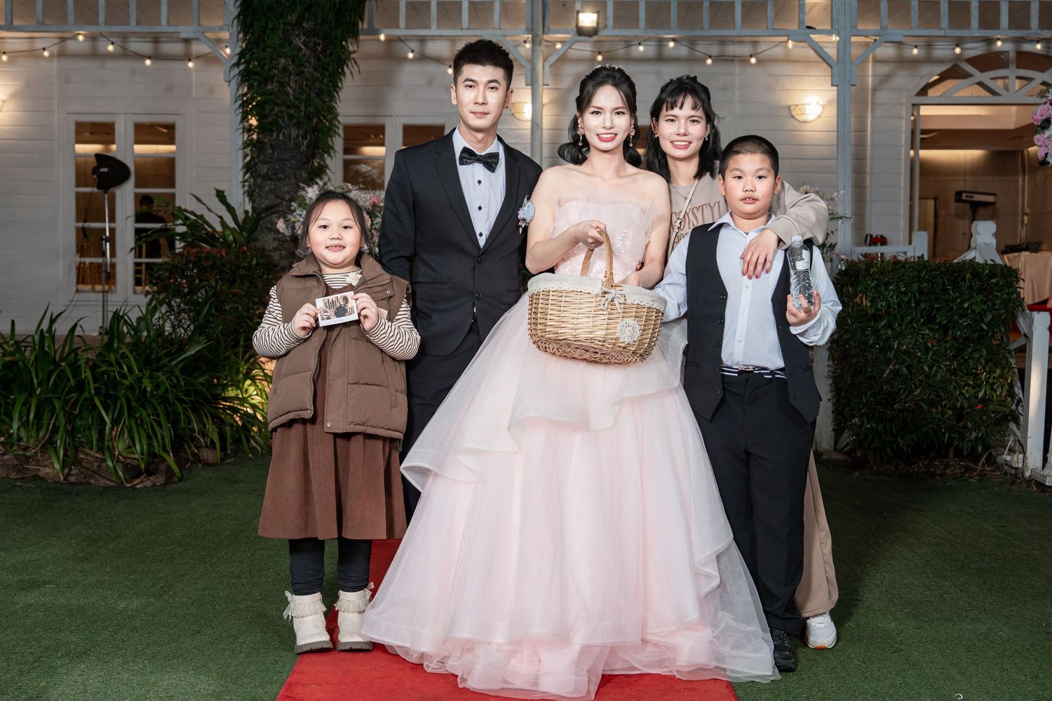 A group of wedding party members in formal attire pose together at an outdoor garden reception venue.