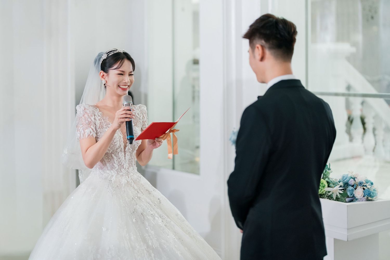 A couple in wedding attire exchanges vows in an elegant white venue with decorative elements.