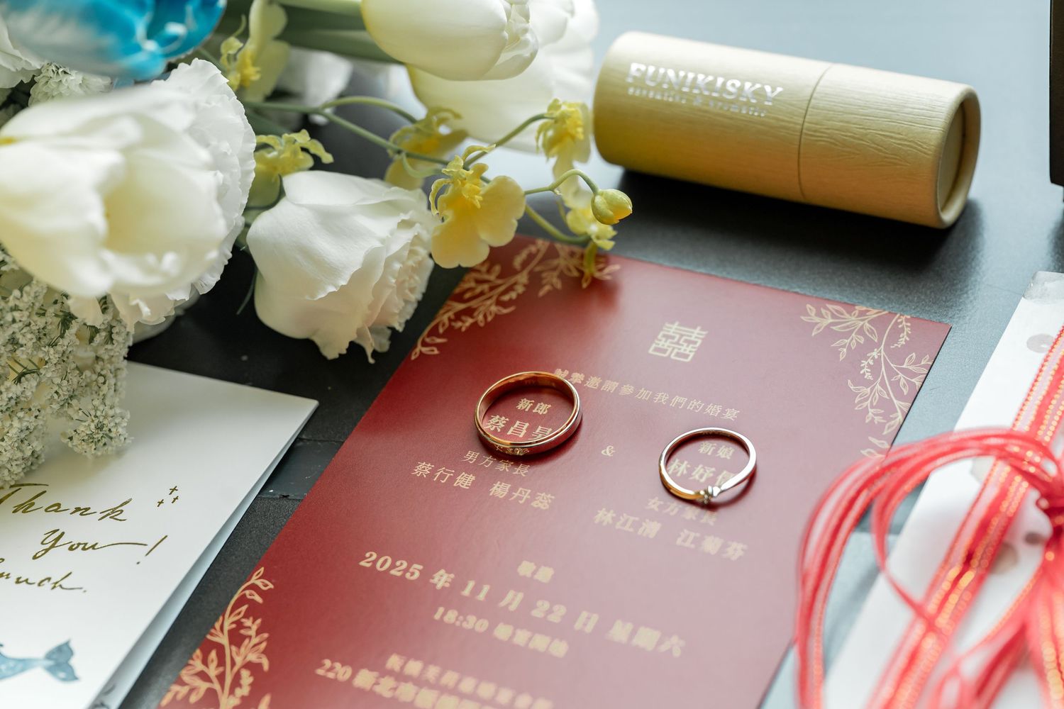 Wedding rings rest on a burgundy invitation card with white floral decorations and golden details.
