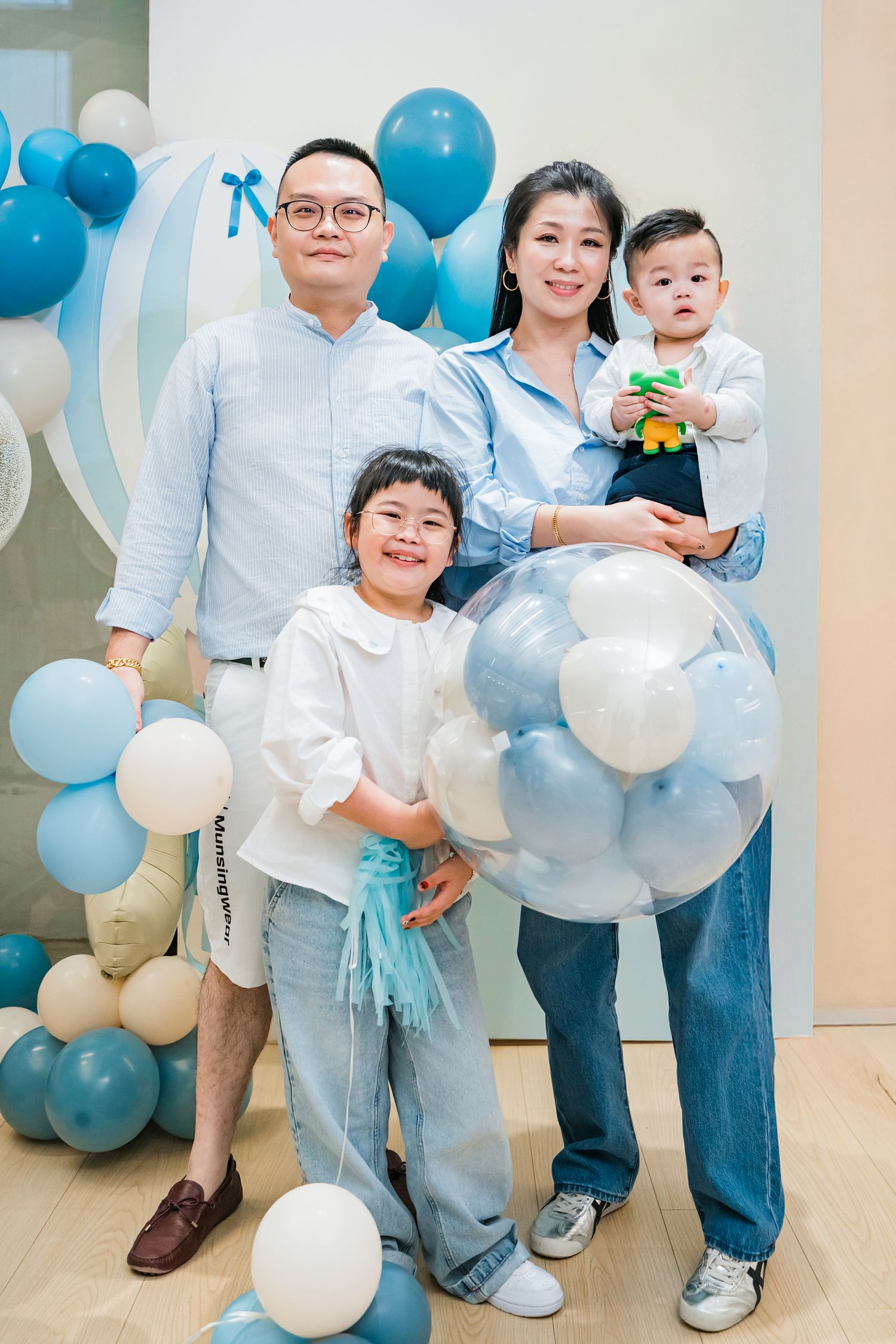 Family poses together at a party decorated with blue and white balloons in a bright indoor setting.