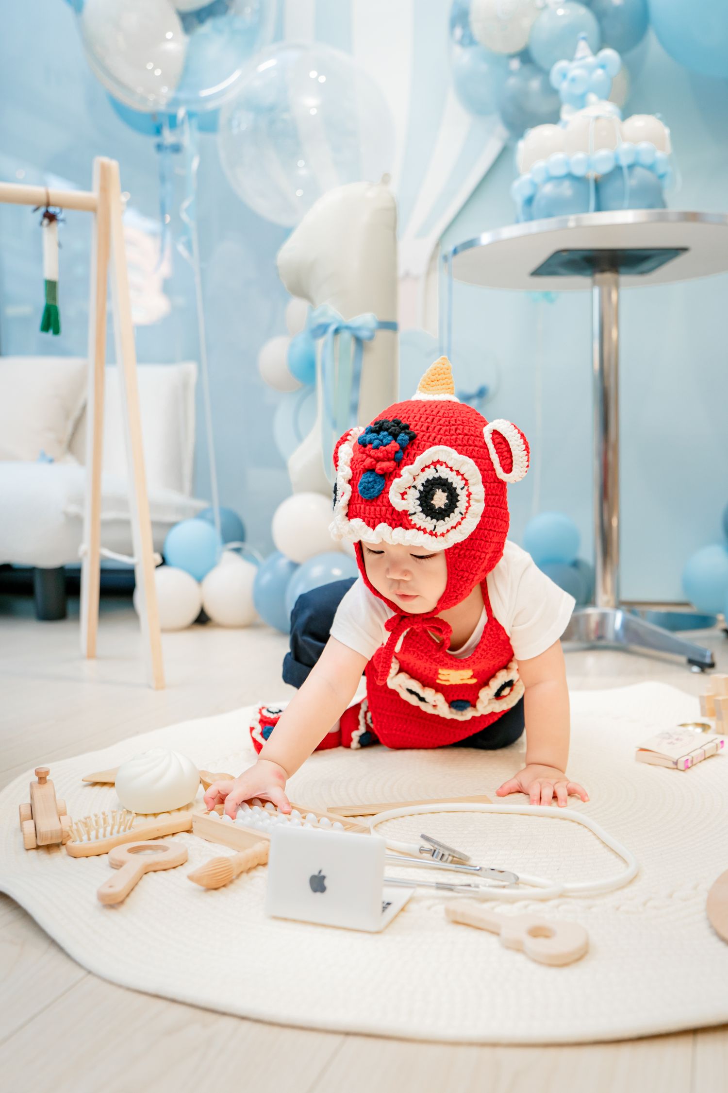 A baby wearing a red knit hat plays on a white mat surrounded by blue balloons in a festive setting.