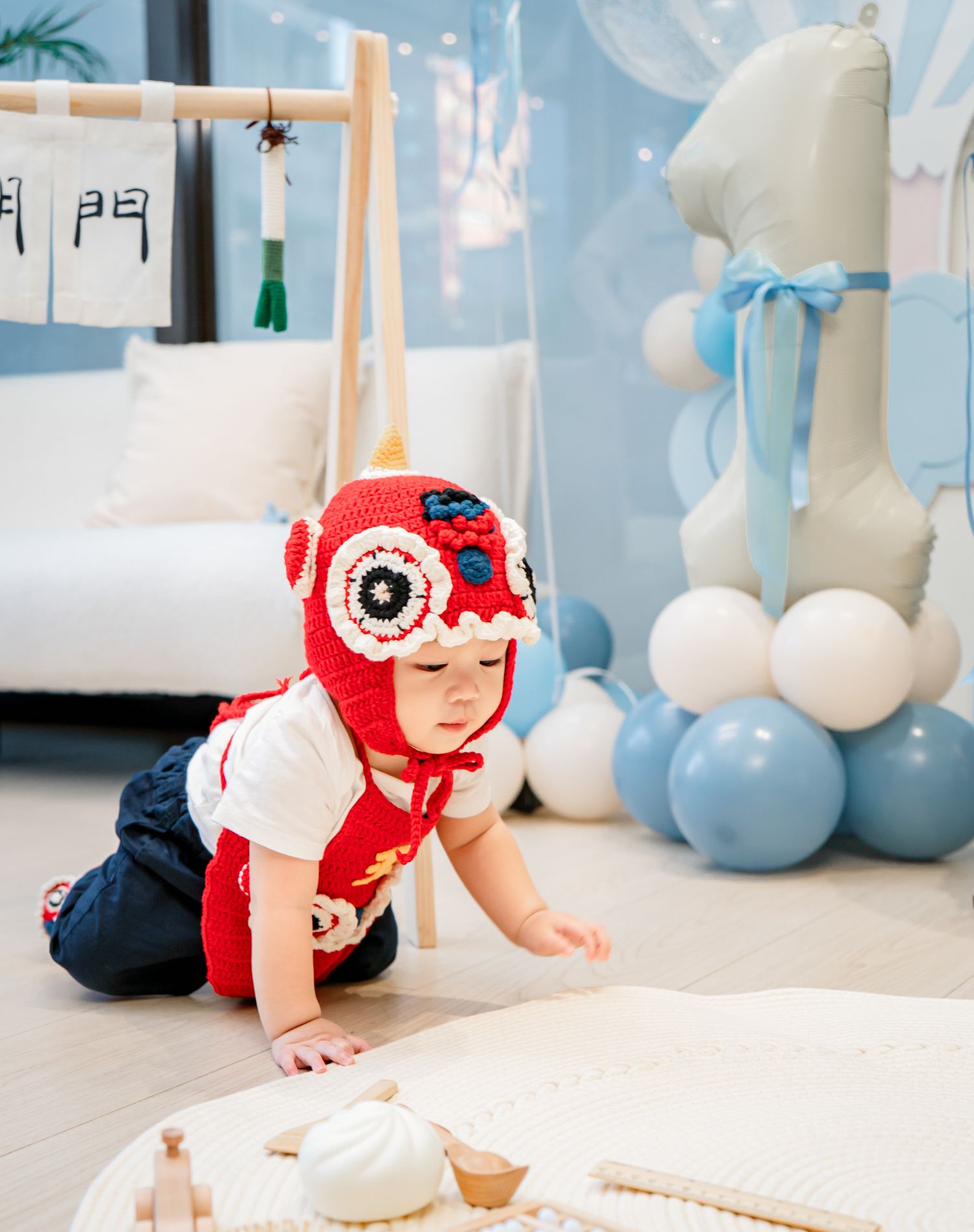 Baby in red knit cap crawls and plays on white mat with blue balloons in background.
