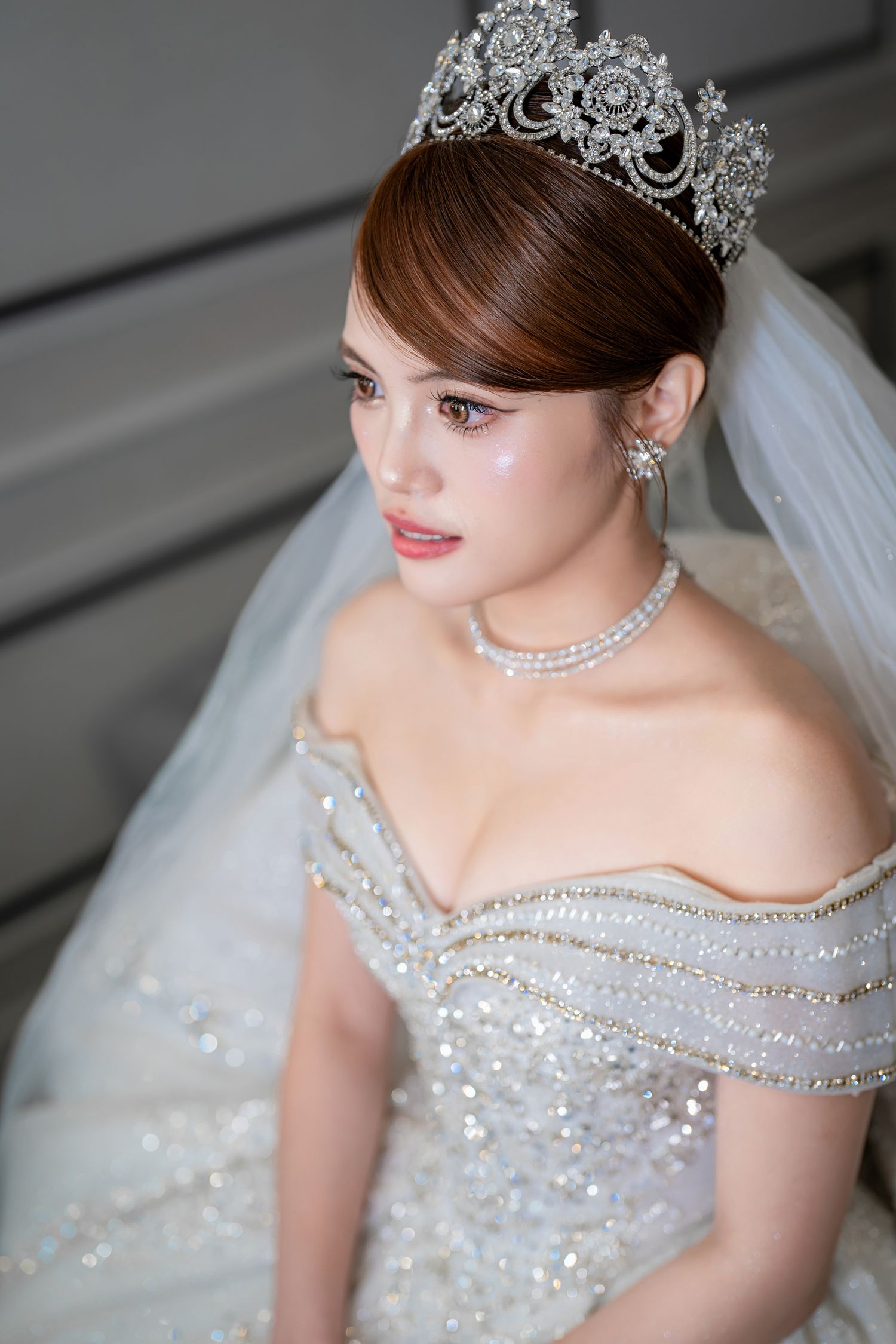 Elegant bride wearing a sparkling crystal tiara, pearl necklace, and off-shoulder beaded wedding gown with a flowing veil.