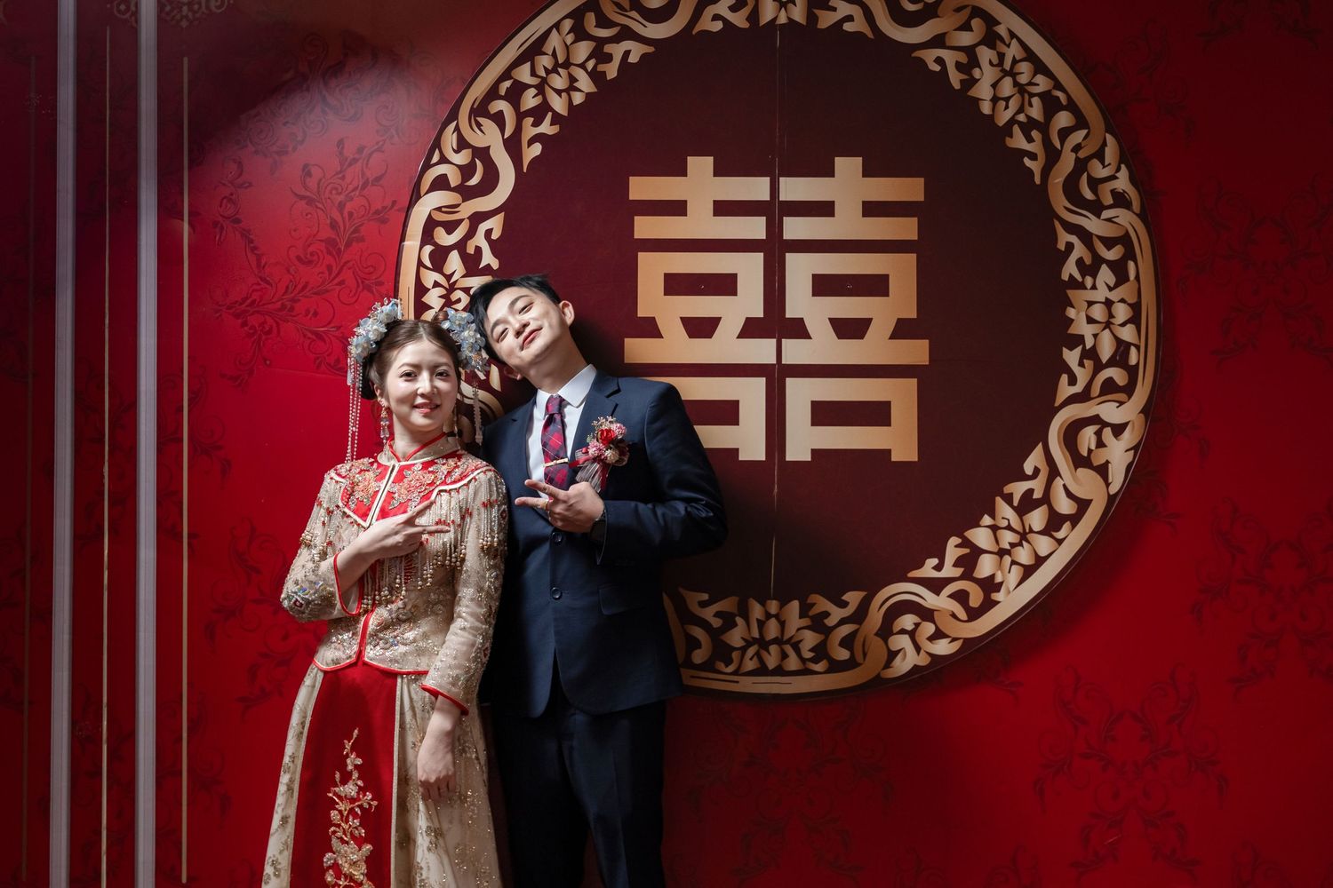 Bride and groom in traditional Chinese wedding attire pose before a red wall with a gold double happiness symbol.