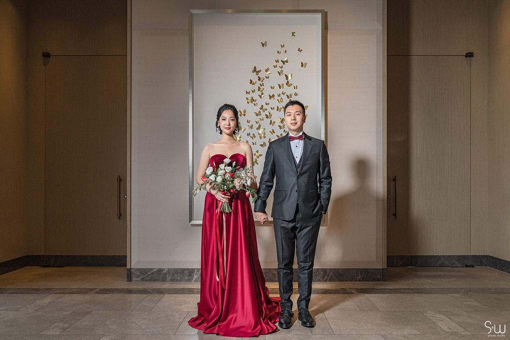 A couple poses formally in front of a butterfly wall display, bride in red gown and groom in black tux.