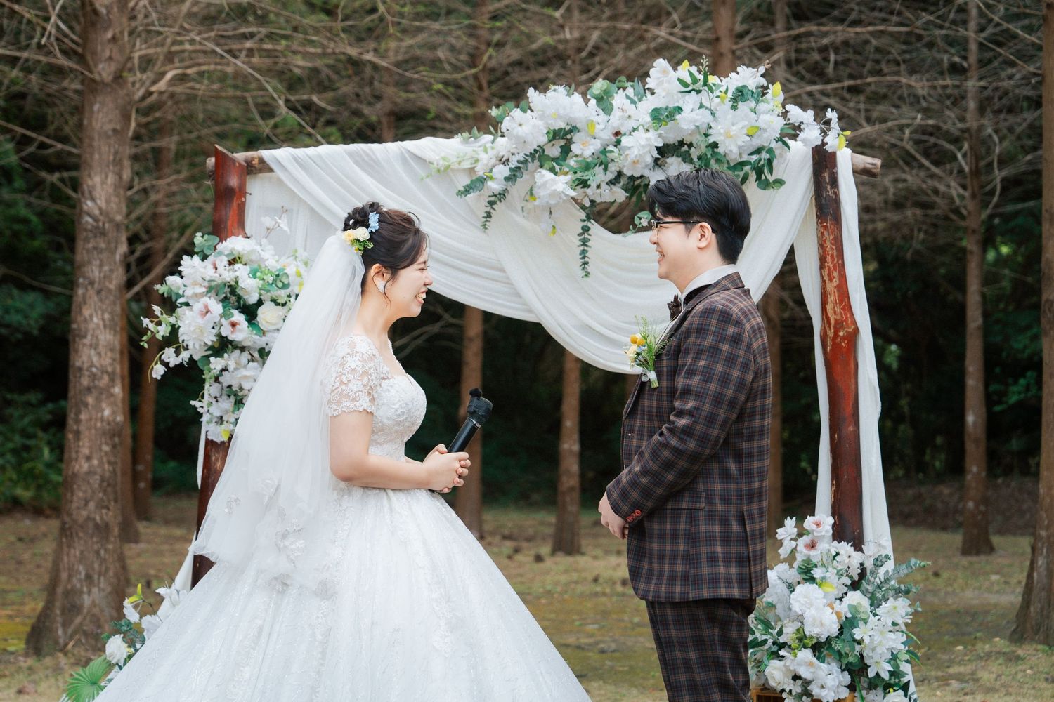 Outdoor wedding ceremony setup with white floral arch and wooden chairs surrounded by evergreen trees.