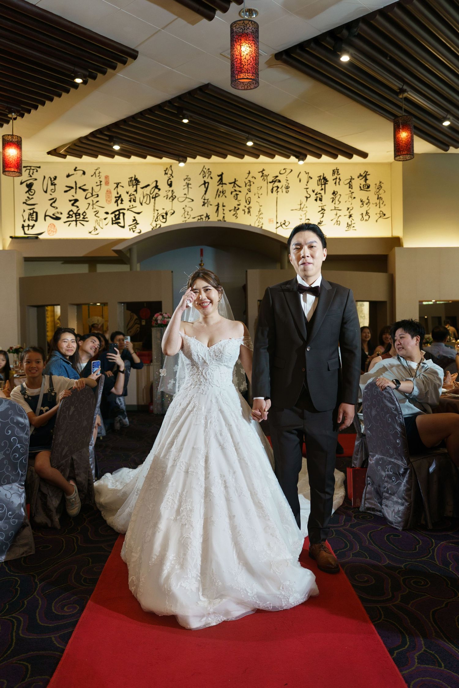 Wedding ceremony in an Asian-inspired venue with red lanterns and calligraphy wall art, featuring a couple on red carpet.