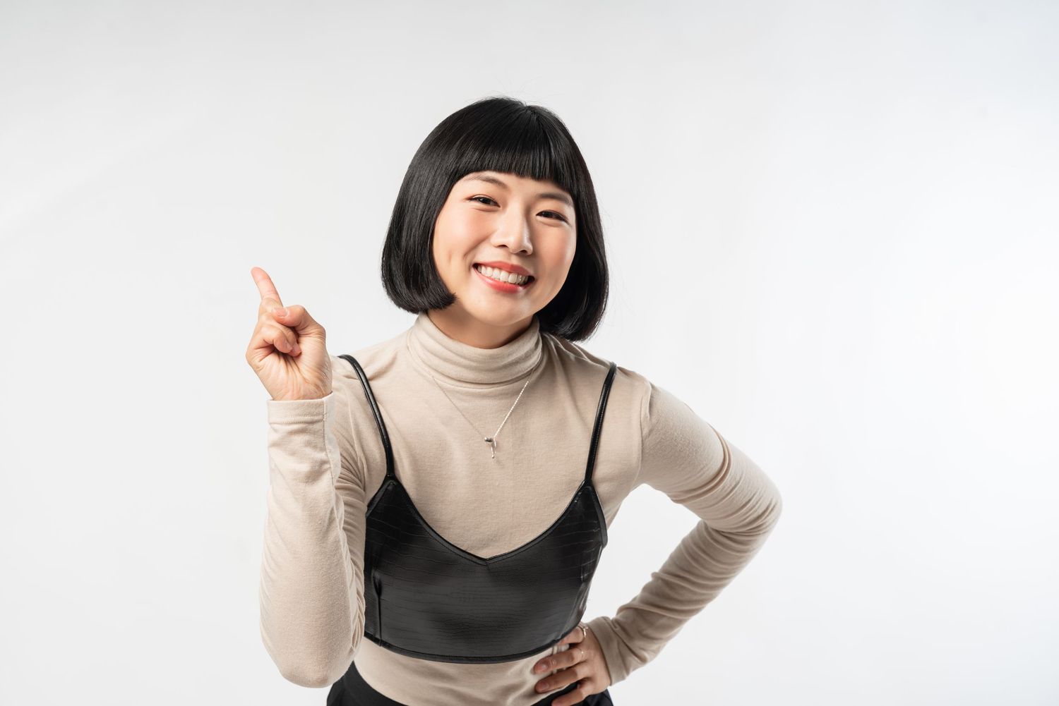 Cheerful person in beige turtleneck and black tank top pointing upward against white background.