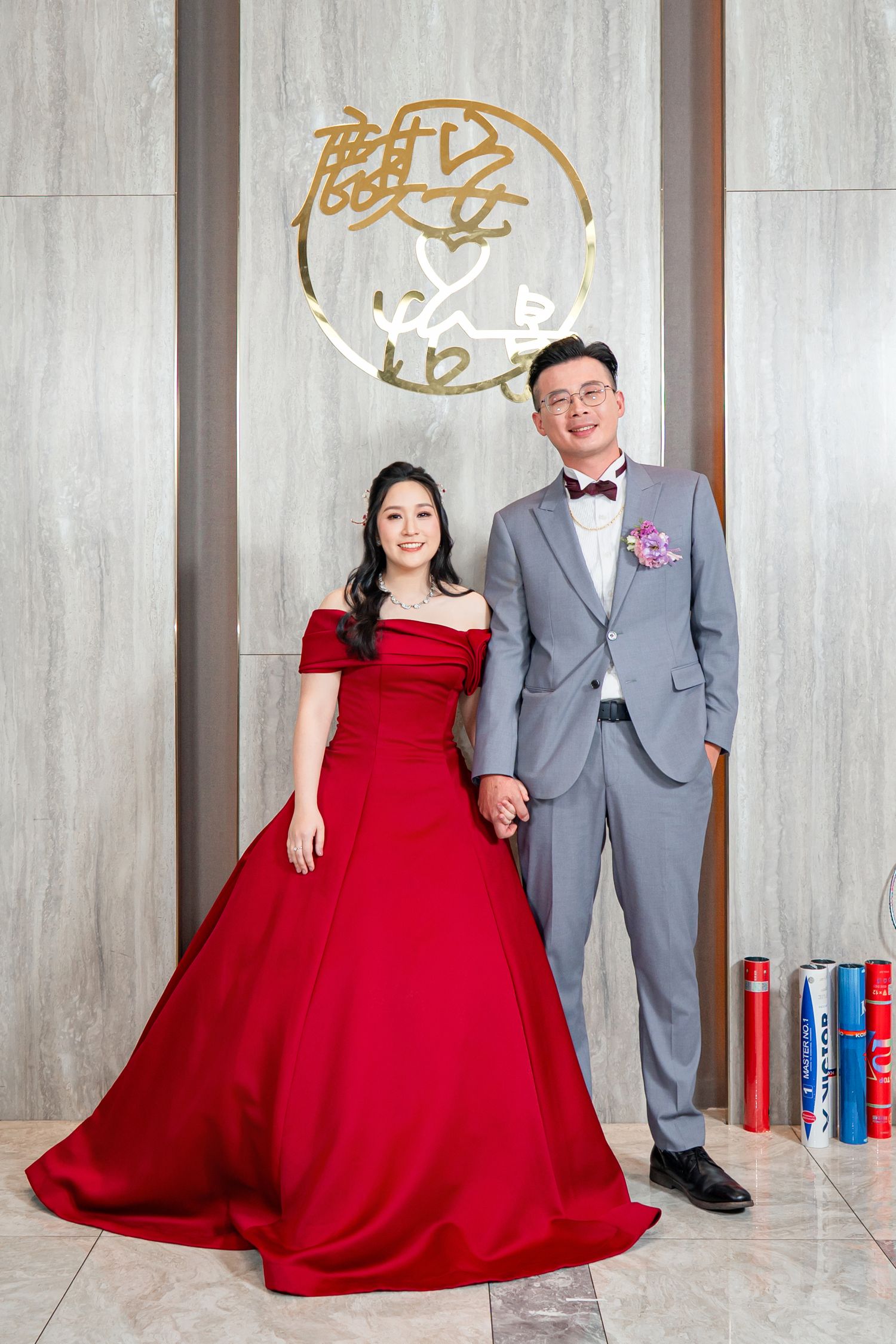 A couple poses at a wedding celebration with the bride in a red off-shoulder ballgown and groom in a grey suit.