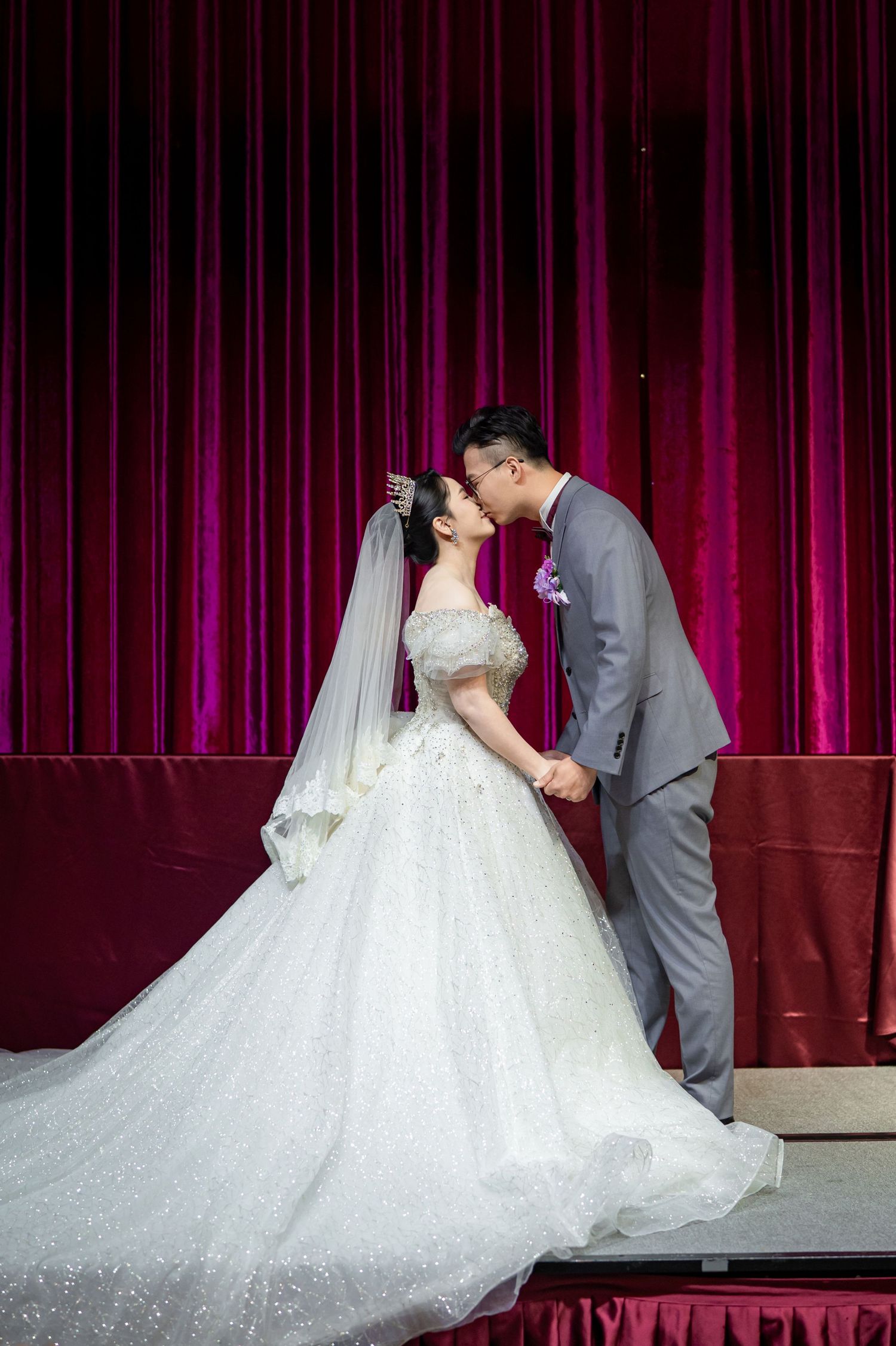 Wedding couple embraces in front of deep red velvet curtain with bride wearing sparkly off-shoulder ball gown and long veil.