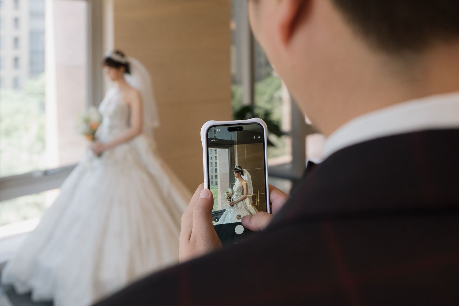 Groom photographing bride in white wedding dress holding bouquet on smartphone at indoor wedding venue.