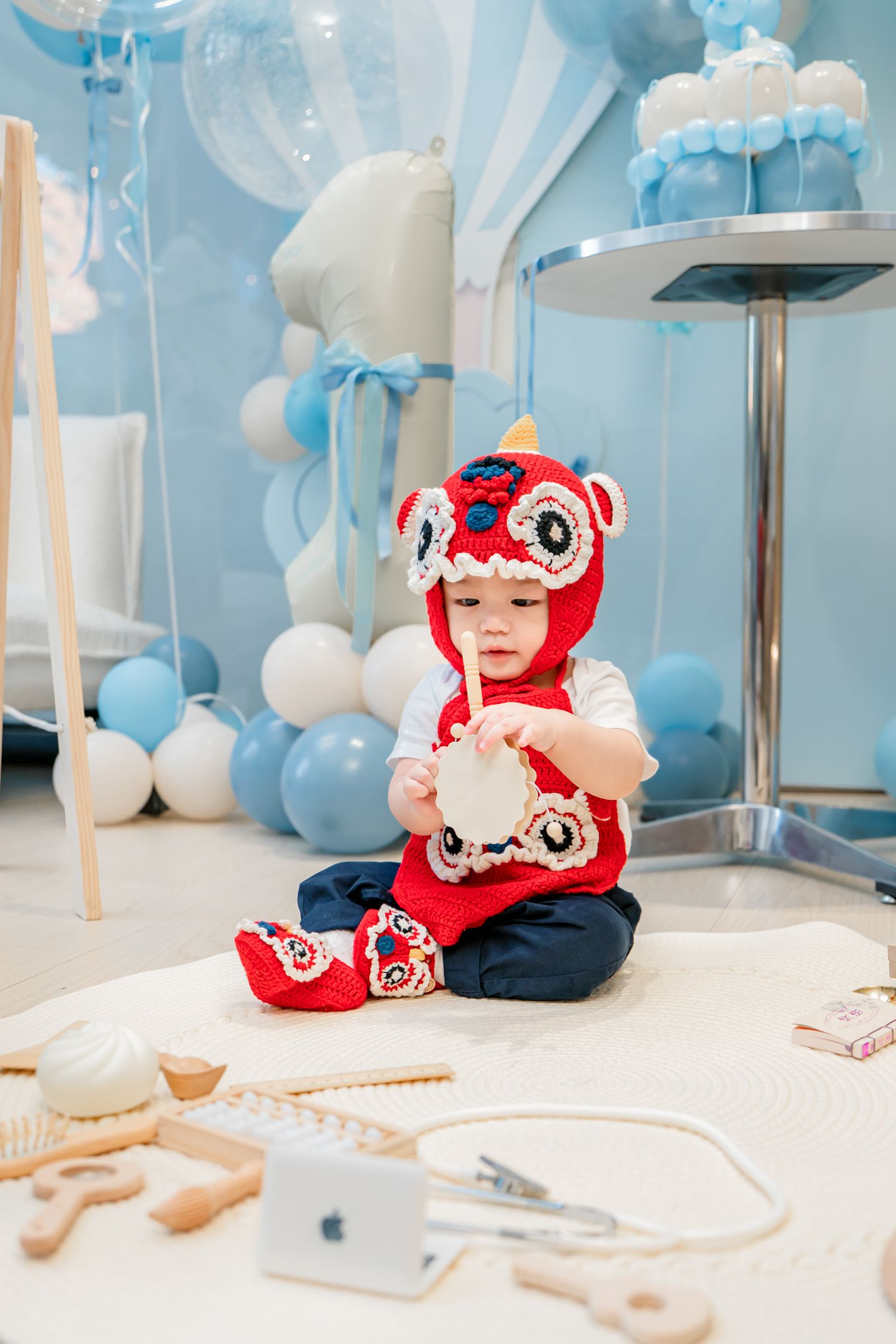 Baby in red daruma doll hat sits at table with blue balloon decorations and party supplies in background.