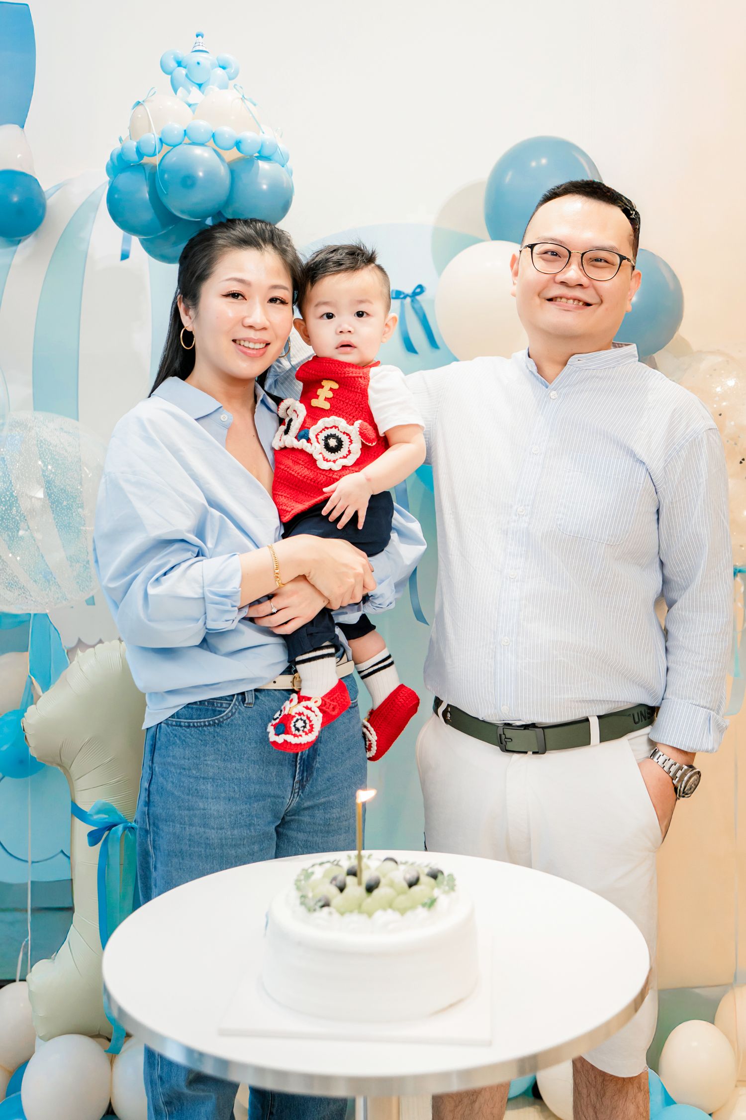 Family celebrates a birthday party with blue and white balloons, cake and matching outfits in a bright indoor setting.